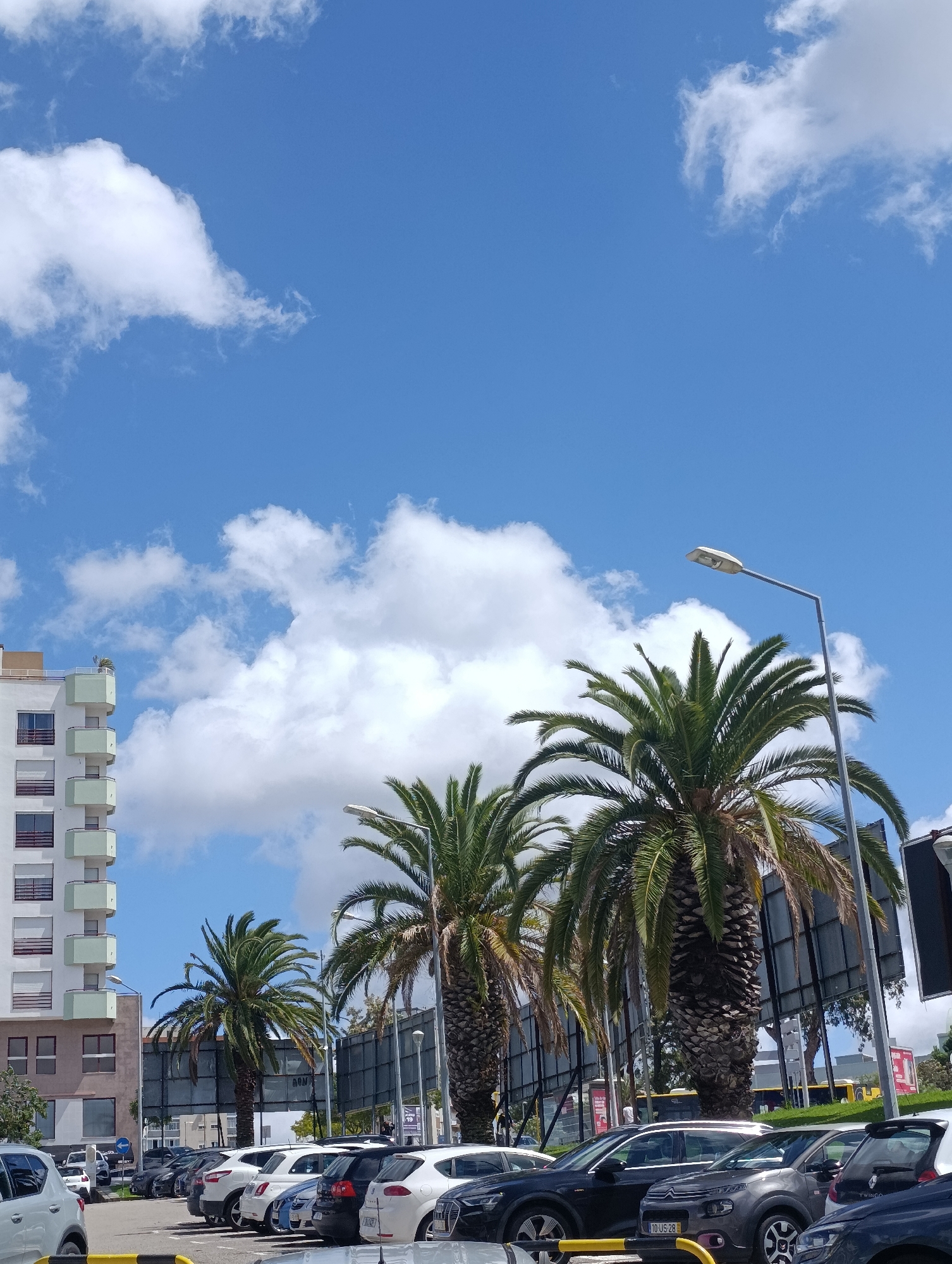 The image shows a parking lot with several cars and palm trees under a blue sky with white clouds. A modern white building with light green balconies is visible on the left side of the frame. A street lamp stands near a tall palm tree in the center-right. The parking lot appears to be in a city, with other buildings and structures visible in the background. The scene is well-lit, suggesting it is daytime. This image was captured in Setúbal, Portugal, specifically in the vicinity of the Parque do Bonfim.