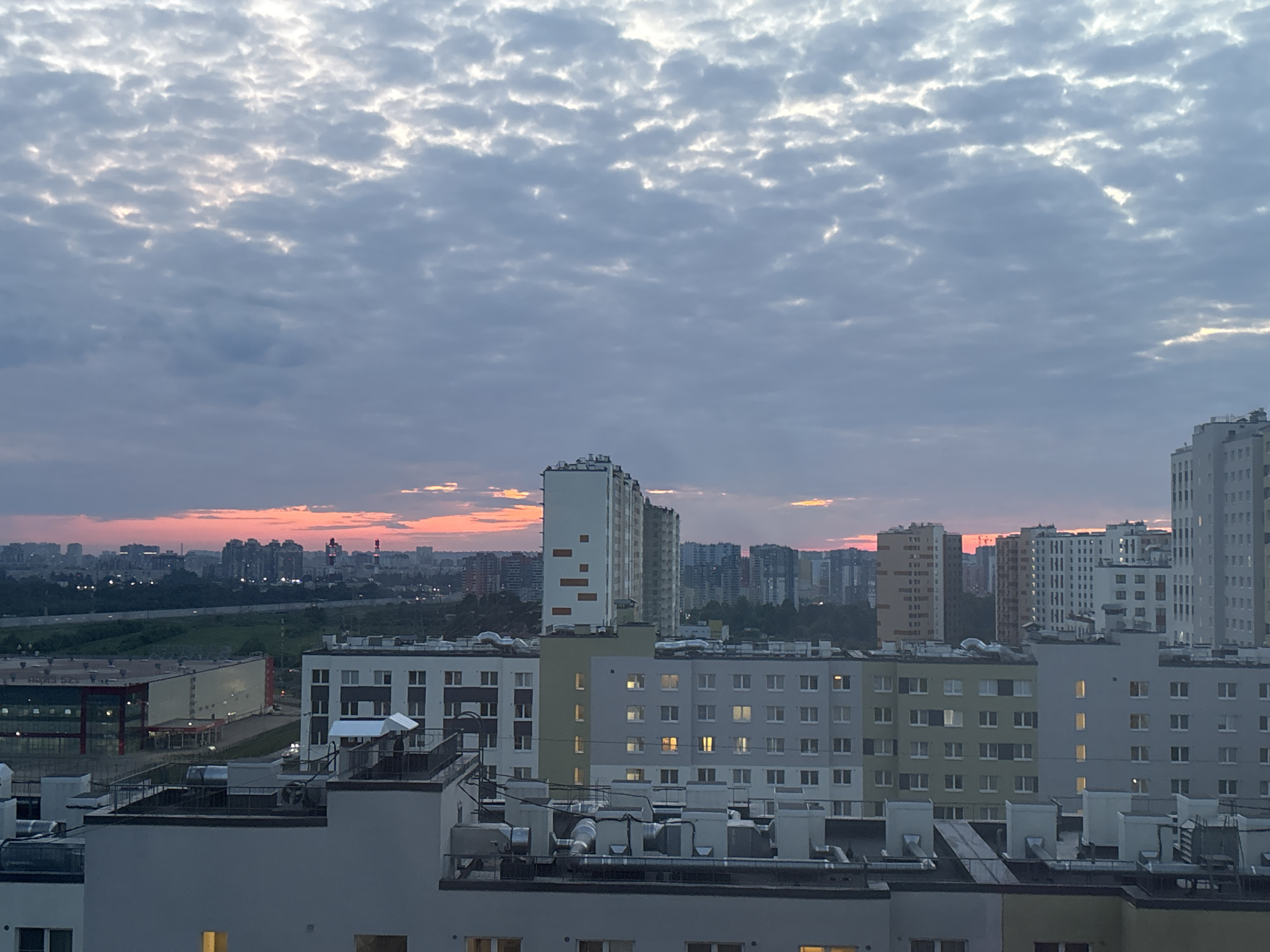 The image provides a wide, elevated view of an urban or suburban landscape at dusk, likely in Vsevolozhsk, Russia. The sky is a prominent feature, almost entirely covered by a dense layer of small, lumpy gray clouds, characteristic of altocumulus or stratocumulus. Patches of lighter, brighter clouds are visible higher up, suggesting diffuse light breaking through. Along the horizon, particularly in the left and central portions of the frame, a distinct band of orange and pink light indicates the setting sun, casting a soft, warm glow.
The scene is dominated by numerous buildings. In the mid-ground, several modern multi-story residential blocks are visible, primarily in shades of light gray and white, featuring many rectangular windows. Some windows display warm interior lights, suggesting evening occupation. A tall, slender white high-rise building with unique rectangular orange-beige accents stands out centrally. To its left, a large, low-rise industrial or commercial structure with a light gray facade and red structural framework is visible, bordered by a green area and what appears to be a road or highway.
Further in the distance, a sprawling cityscape extends to the horizon, with countless high-rise buildings silhouetted against the colorful sky. The foreground shows the rooftops of closer buildings, featuring various ventilation units, pipes, and air conditioning systems. No people are visible in the scene. There is no discernible text present on any of the buildings or signs. The overall atmosphere is calm and tranquil, capturing the transition from day to night over a busy urban environment.