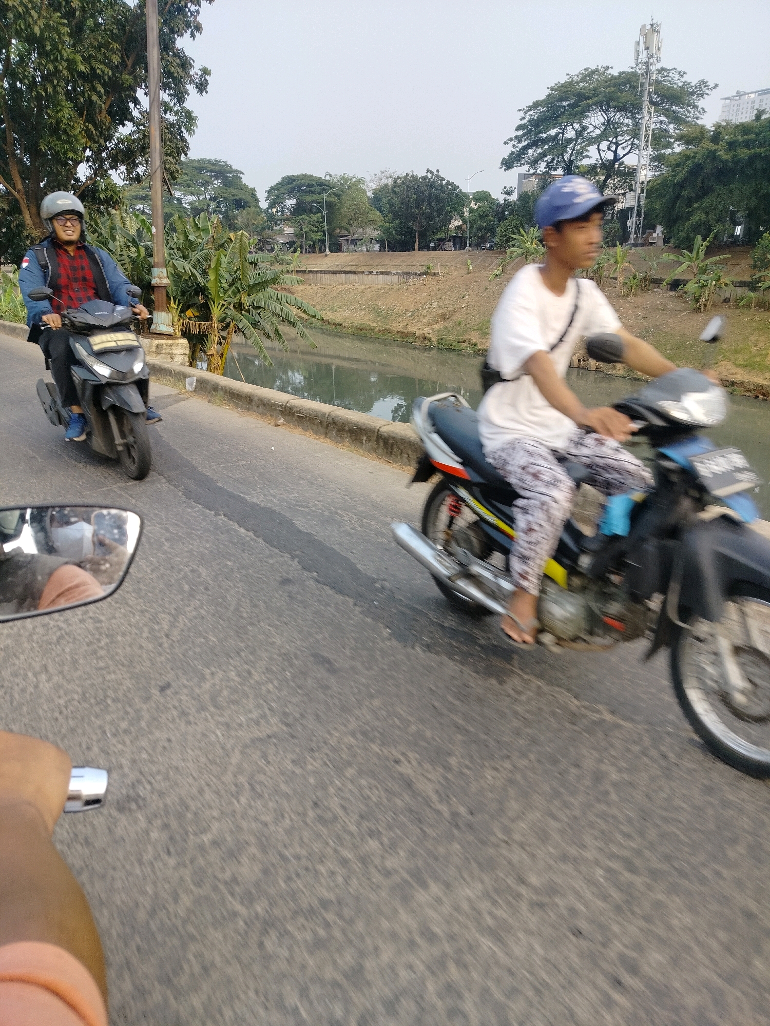 Two individuals are riding motorcycles on a paved road next to a canal. The person in the foreground, on the left, is wearing a blue jacket over a red and black checkered shirt, black pants, blue shoes, and a grey helmet. They are riding a black scooter. The person in the background, on the right, is wearing a white t-shirt, patterned pants, and a blue baseball cap. They are riding a blue and black motorcycle. The canal water is murky, and the banks are lined with trees and vegetation. In the distance, a cell phone tower and some buildings are visible, suggesting an urban or semi-urban environment. The sky is overcast. The scene is captured from a low angle, likely from another moving vehicle, as indicated by the blurry foreground and the reflection in a mirror. This scene is consistent with the landscape of East Jakarta, Indonesia, which features numerous canals and urban development alongside natural elements.