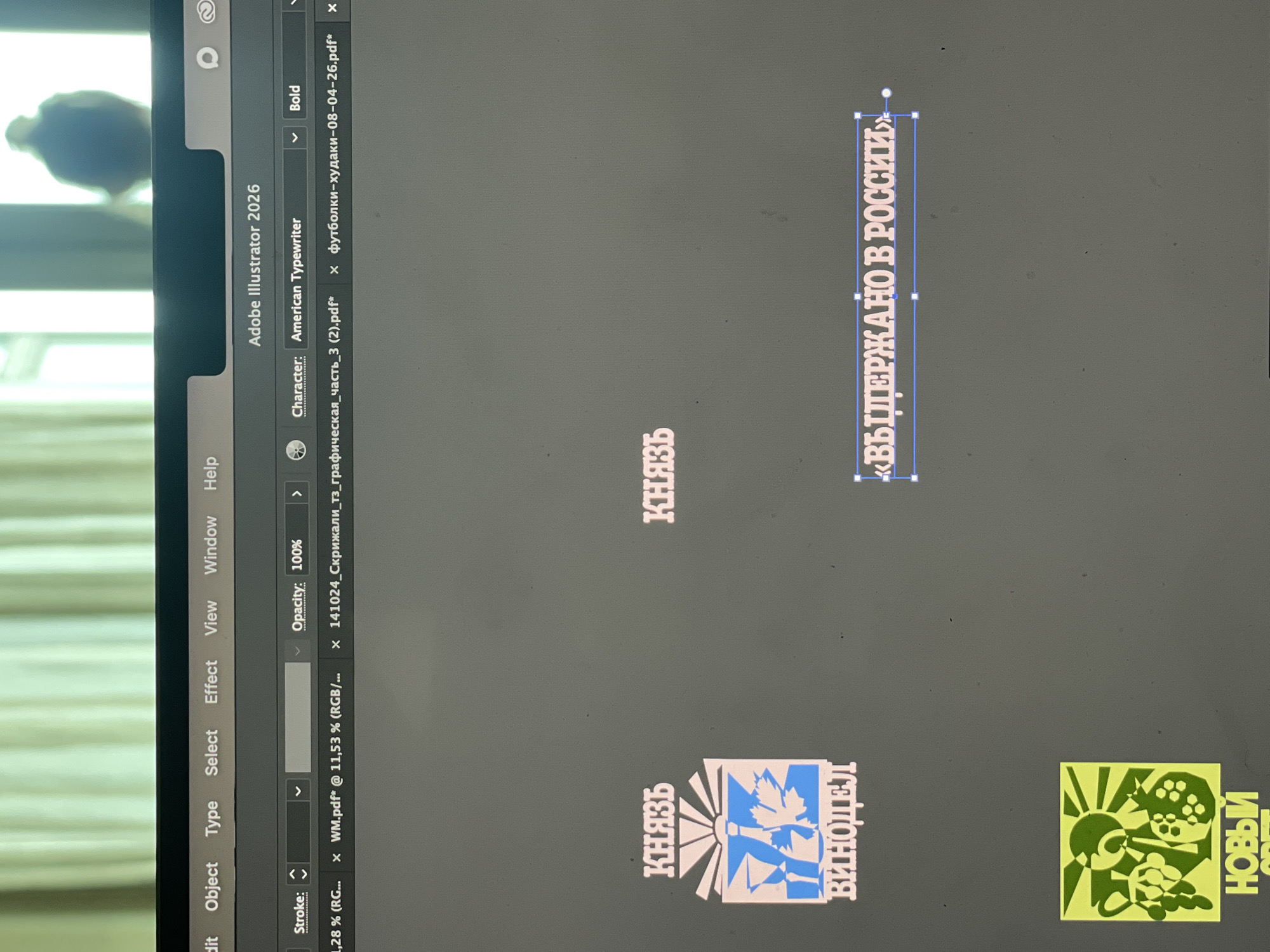 The image displays a computer screen, likely a laptop or desktop monitor, showing Adobe Illustrator 2026 in use, set against a blurred, out-of-focus indoor background in Bangkok, Thailand. The background suggests an interior space with natural light entering from a window, and a dark, indistinct shape is faintly visible in the upper left corner.
On the screen, the Adobe Illustrator interface is visible with its menu bar (including "Edit," "Object," "Type," "Select," "Effect," "View," "Window," "Help"), a character panel indicating "American Typewriter" font in "Bold," and an opacity setting of "100%." Several file tabs are open, including "WM.pdf*", "141024_Скрижали_тз_графическая_часть_3 (2).pdf*", and "футболки-худаки-08-04-26.pdf*".
The main area of the screen shows a dark gray design canvas populated with several graphic elements, predominantly featuring Cyrillic text.
Visible text includes:
* "КНЯЗЬ" (KNYAZ, meaning "Prince" or "Duke"), appearing twice independently.
* "ВИНОДЕЛ" (VINODEL, meaning "Winemaker").
* "«ВЫДЕРЖАНО В РОССИИ»" ("VYDERZHANO V ROSSII," meaning "Aged in Russia" or "Produced in Russia"), which is highlighted with a blue selection bounding box.
* Partially visible text "НОВЫЙ" (NOVY, meaning "New") and "СВЕТ" (SVET, meaning "Light" or "World") as part of a logo.
Two distinct logo-like graphics are present:
1. A white and blue design featuring a stylized sunburst, with the text "КНЯЗЬ" and "ВИНОДЕЛ" integrated vertically.
2. A green and yellow stylized sunburst design with abstract shapes, accompanied by the partially visible text "НОВЫЙ СВЕТ."
The activity taking place is graphic design or document review within Adobe Illustrator, likely involving branding or packaging design given the logos and text about "Prince," "Winemaker," and "Aged in Russia." The light from the screen illuminates the immediate area, and the blurred background implies a bright daytime environment indoors.
