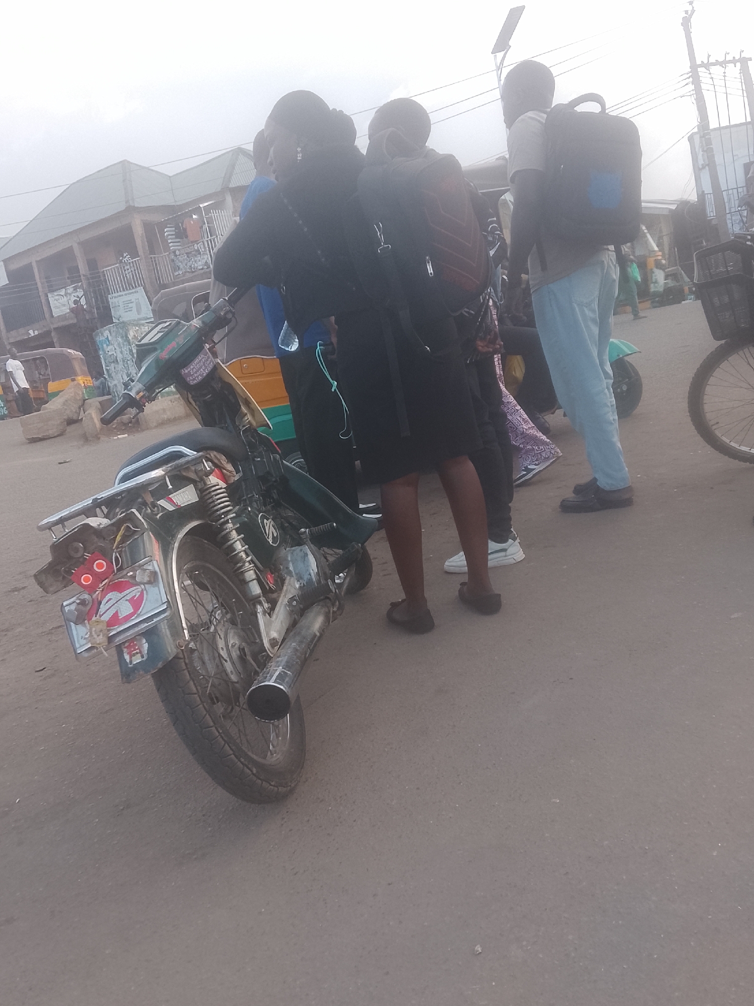 The image depicts a street scene in Gwong, Nigeria. In the foreground, a green Yamaha motorcycle with a damaged rear light and exhaust pipe is parked. Several people are standing nearby; they wear backpacks and casual clothing, suggesting they are students or commuters. In the background are buildings, other motorcycles, and a bicycle. The setting is a busy street with visible signs of urban activity. The scene conveys a typical day in an urban area, with people utilizing public transportation and the visible signs of wear and tear on the vehicles and environment.