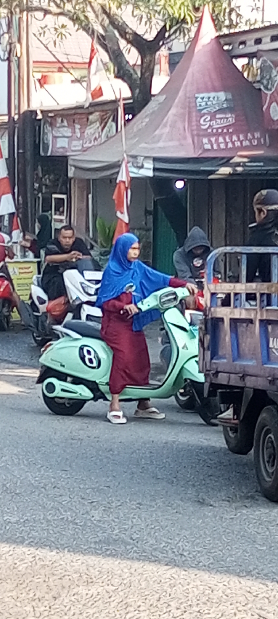 In Setu, Indonesia, a woman wearing a blue hijab and a maroon dress is riding a light green scooter. She is holding the handlebars and looking forward. The scooter has the number "8" in a black circle on its side. Behind her, a man in a black shirt and pants is sitting on a white scooter. To the right of the woman, a man in a grey hooded sweatshirt is partially visible. A blue truck is on the right side of the image, with its rear end prominent. Red and white Indonesian flags are visible in the background, along with a red tent with text on it. The scene appears to be on a paved street with trees and buildings in the background.