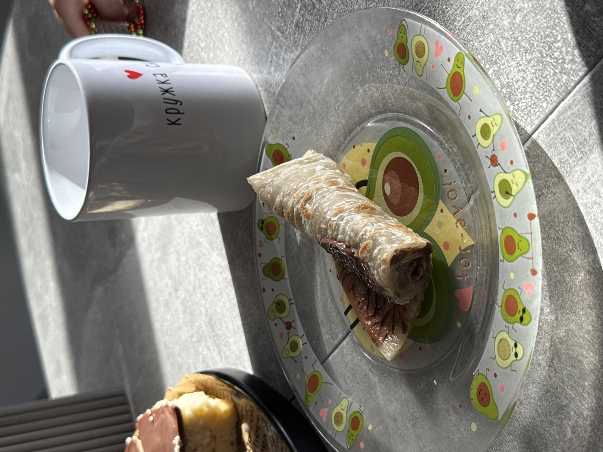 An overhead shot shows a rolled crepe filled with chocolate spread on a clear glass plate decorated with avocados. To the right, a white mug with the Russian inscription "кружка с" and a red heart sits on a gray tiled surface. In the background, a fluted gray object is visible on the left. The scene is lit by harsh sunlight, casting strong shadows. The location is stated to be Bortuki, Russia.