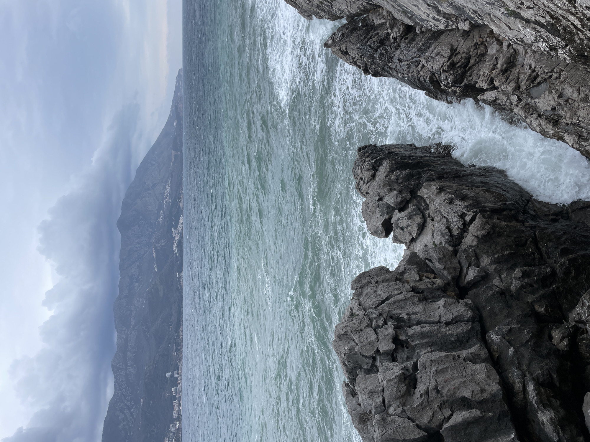 The image displays a turbulent coastal scene in Budva, Montenegro. The foreground is dominated by rugged, dark grey rock formations, exhibiting sharp edges and deep fissures. Waves crash against these rocks, generating significant white foam, particularly within a narrow channel between two large rock masses on the right. The midground features a vast expanse of choppy, dark teal-green seawater, characterized by numerous whitecaps and discernible swells. In the background, a range of dark green mountains spans the horizon. Scattered white and light-colored buildings, indicative of coastal settlements, are visible along the base and lower slopes of these mountains, particularly on the left. The sky is heavily overcast with dense, dark grey clouds. No individuals or specific man-made objects, aside from the distant structures, are present.