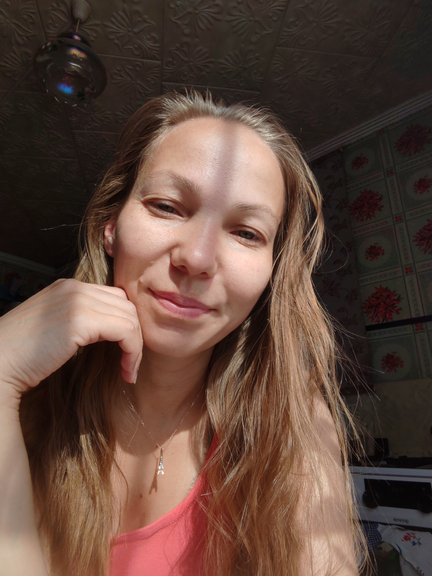 A close-up, high-angle, selfie-style photograph shows a young Caucasian woman with long, light brown hair and fair skin. She is looking directly at the camera with a slight smile, resting her chin on her left hand. A thin, gold necklace with a small Eiffel Tower pendant is visible around her neck. She is wearing a coral-pink sleeveless top. The background features a textured ceiling with embossed floral patterns and floral-patterned wallpaper. A ceiling light fixture with a glass shade is visible in the upper left corner. Sunlight casts shadows across her face and hair, indicating it is daytime. The environment appears to be an indoor residential setting, possibly a kitchen based on the edge of a stove visible in the lower right, suggesting the location is within the city of Chukhloma, Russia.