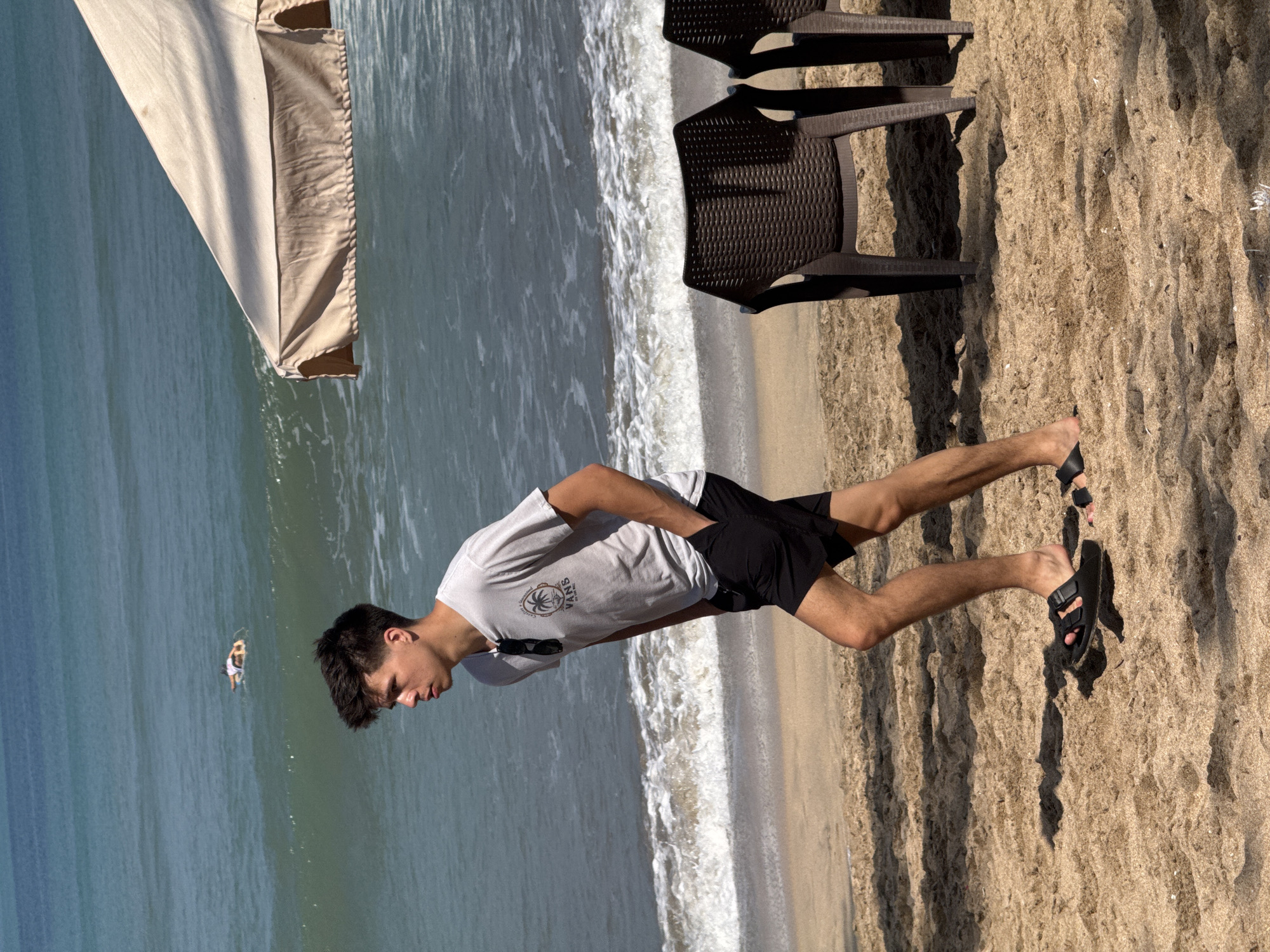 A young man with dark hair, wearing a light gray t-shirt with a palm tree graphic and black shorts, walks on a sandy beach. He has sunglasses hanging from his t-shirt collar and is wearing black sandals. The beach is beside a body of water with small waves breaking on the shore. In the distance, a person is visible in the water on a floating device. Two dark plastic chairs are placed on the sand to the right of the frame, under a beige umbrella. The scene appears to be a public beach in Kuta, Indonesia.