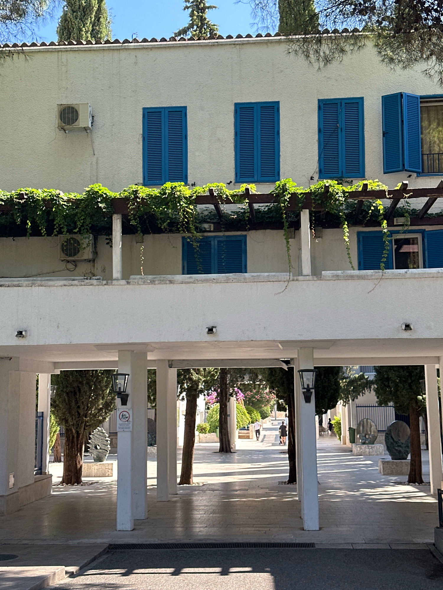 The image shows a white, multi-story building with blue shutters on its windows. A pergola covered in green ivy runs along the second floor, supported by white pillars. Below this, a covered walkway, also supported by white pillars, leads into an outdoor area with trees, sculptures, and people walking. The walkway has a paved floor and is lit by lanterns. In the background, more trees and foliage are visible, suggesting a sunny day.
The location in the city of Budva, Montenegro is the Modern Gallery (Moderna Galerija Budva). The sculptures visible in the background are part of the gallery's outdoor exhibition.