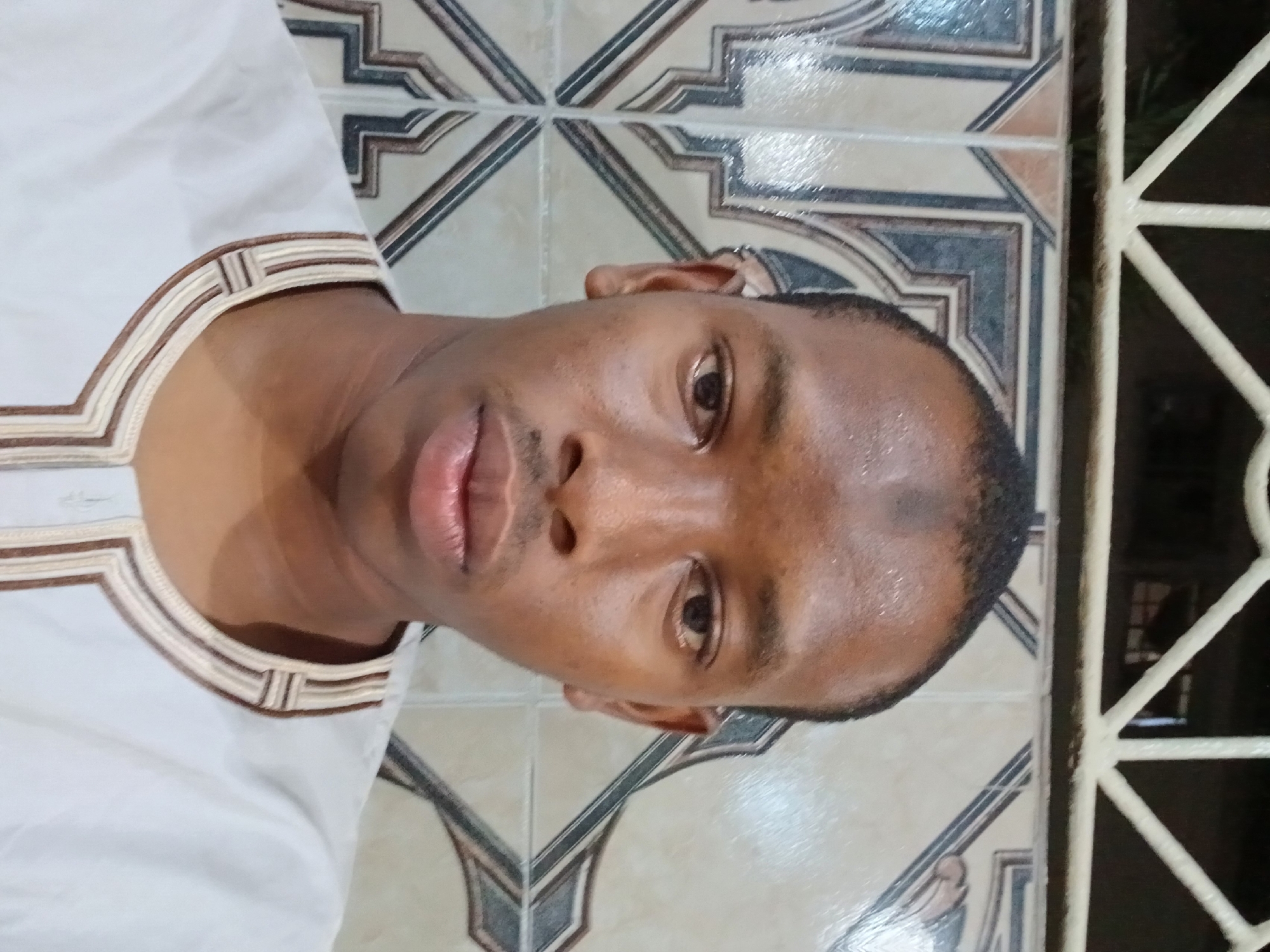A close-up shot shows a man in a white shirt with brown trim around the collar, positioned in front of a tiled wall with a geometric pattern. To the right of the man, a white barred structure is partially visible, suggesting a window or partition. The lighting is diffuse and the man's skin appears slightly shiny, indicating possible perspiration. Without further details, the specific location in Lagos, Nigeria cannot be determined.