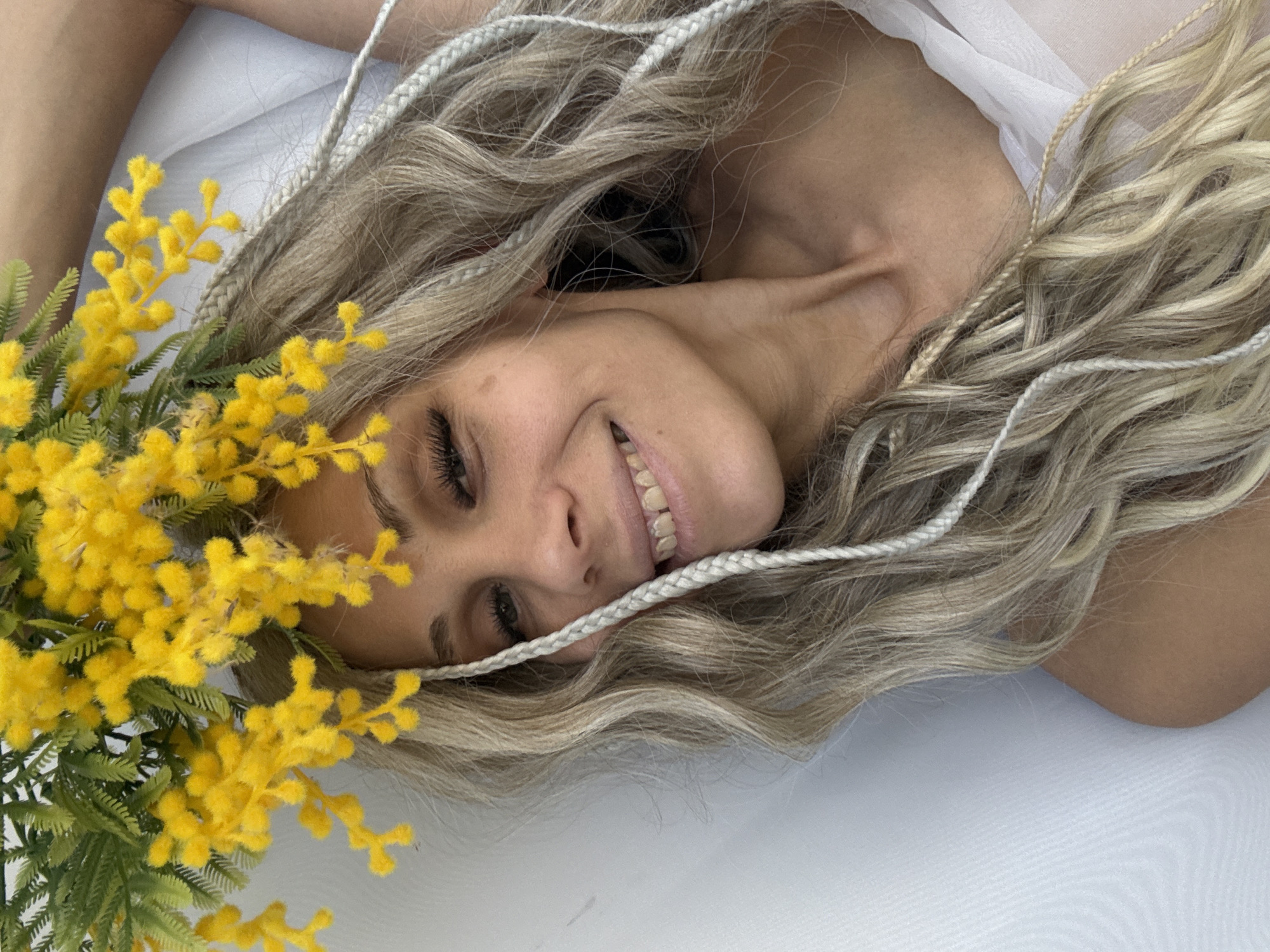 A woman with blonde hair with light-colored braids interwoven throughout is lying down on a white surface. She is smiling with yellow flowers near her head. She is wearing a white top. The location is unknown.