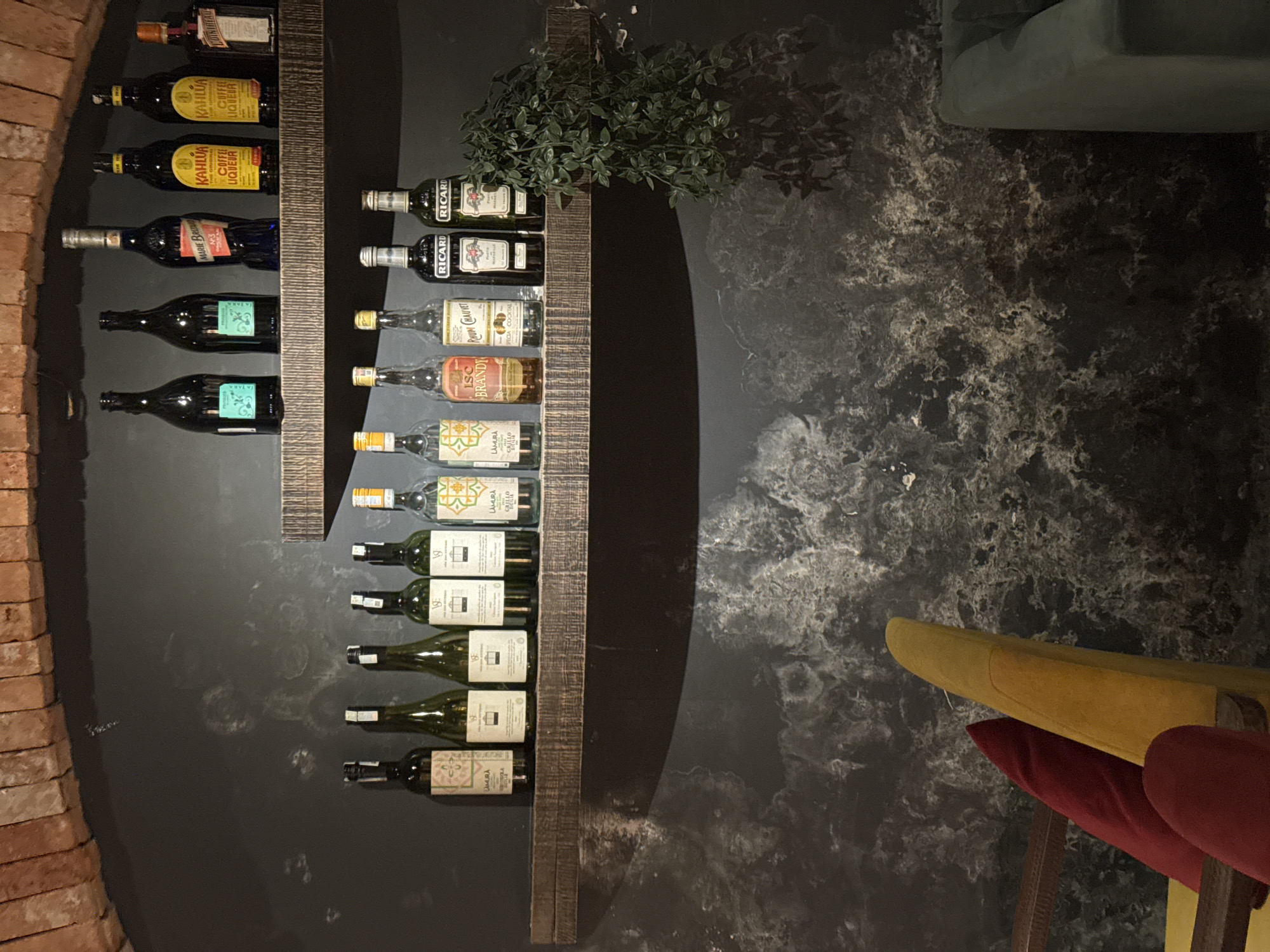 The image shows a well-stocked wine rack against a dark, marbled wall. The rack is made of rough-hewn wood and holds numerous bottles of various spirits and liqueurs. On the left side of the rack, there are bottles of Kahlúa, Marie Brizard, and other dark liquors. On the right side, there are bottles of Ricard, Rhum Gauvauet, and other clear liquors. Some of the visible text on the labels includes "KAHLUA," "COFFEE LIQUEUR," "MARIE BRIZA," "RICARD," "LSC BRANDY," and "RHUM GAUVET."
In the upper portion of the rack, a small artificial plant with green leaves casts a shadow on the wall. The overall lighting is dim, suggesting an indoor setting, possibly a bar or a restaurant. The lower right of the image shows the edge of a yellow and red upholstered chair, hinting at seating for patrons. A brick arch is visible on the far left of the image, adding to the architectural details of the space. The scene appears to be static, with no people or ongoing activity depicted.