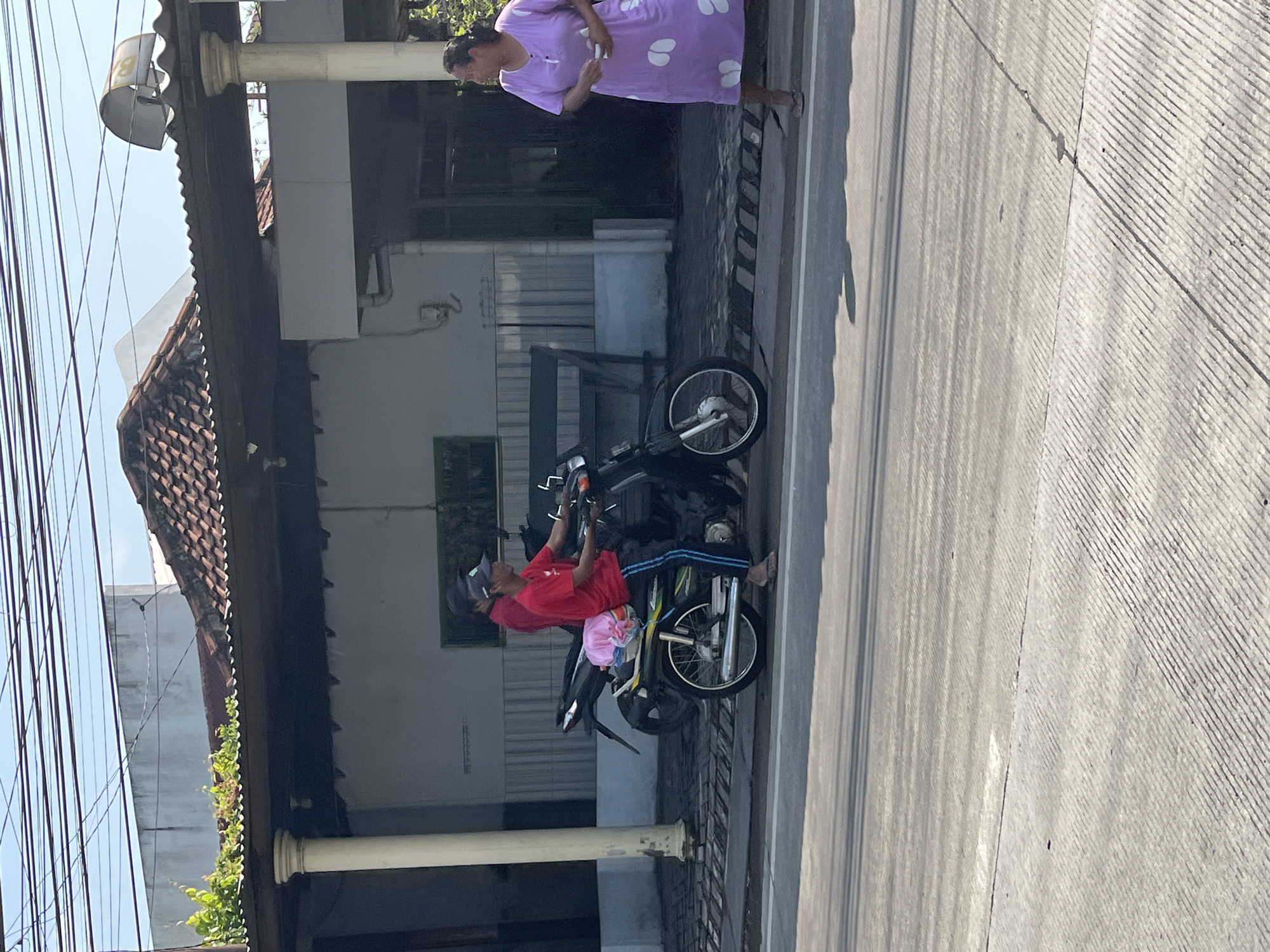 This image depicts a street scene in Kepadangan, Indonesia. A man in a red shirt and black pants is riding a motorcycle, with a second person, also in a red shirt, seated behind him. The motorcycle has a pink bag on the back. To the right, a woman in a purple dress with a white pattern is standing on the sidewalk, holding a white object. The background shows a building with a tiled roof and pillars, and power lines are visible overhead. The street in the foreground is paved.