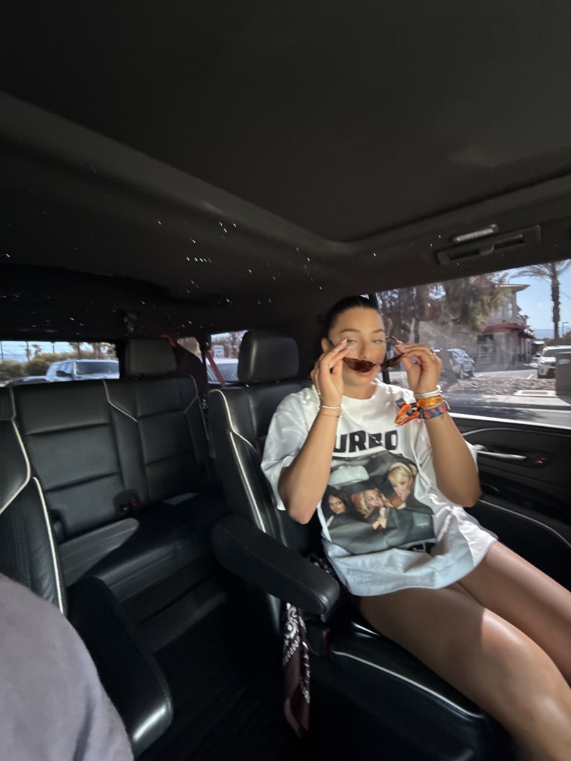 A young woman is captured inside the luxurious interior of a vehicle, likely an SUV or van, during bright daylight. She is seated on a black leather passenger seat with white piping, looking towards the front of the vehicle while adjusting a pair of brown-tinted sunglasses. Her dark hair is pulled back, and she wears a loose-fitting, oversized white t-shirt that features a graphic print. The word "TURBO" is distinctly visible above an illustration of what appears to be three women inside a car on her shirt.
Her bare legs are visible, suggesting she is wearing shorts or a very short skirt. Several bracelets, including some colorful ones, adorn her right wrist. A dark patterned bandana or scarf is casually draped over the armrest beside her. The vehicle's dark ceiling is notable for its integrated small, scattered lights, resembling a "starlight" headliner.
Through the car's windows, blurred scenes of the outside world are visible, showing what appear to be palm trees, buildings, and other vehicles, hinting at a sunny, possibly urban or suburban location. The overall scene suggests a relaxed journey in a high-end car.
