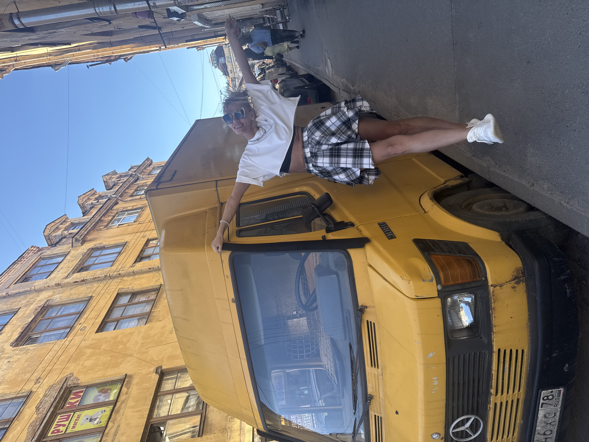 A woman with blonde hair and sunglasses poses with her arms outstretched next to a yellow Mercedes-Benz van parked on a cobblestone street in Saint Petersburg, Russia. The woman is wearing a white cropped t-shirt, a black bra, and a black and white plaid skirt. The van has a license plate that reads "46 XC 78 RUS". The background shows aged yellow buildings with multiple windows and balconies. The sky is clear and blue.