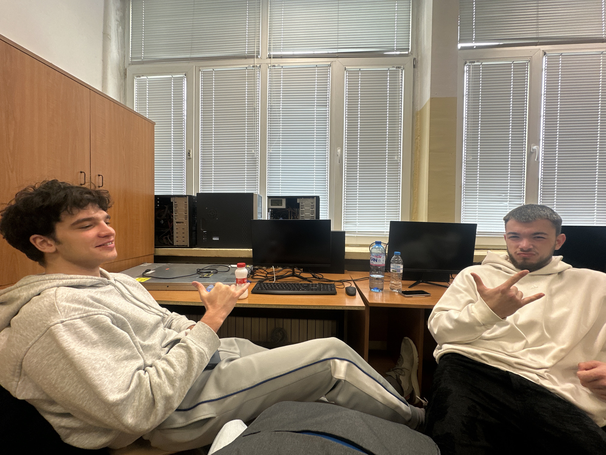 Two young men are seated in an office or classroom setting. The individual on the left has curly dark hair and is wearing a light grey hoodie and sweatpants with a blue stripe. He is making a "shaka" hand gesture. The individual on the right has short dark hair and a beard, wearing a white hoodie and black pants. He is making a hand gesture resembling "rock on" or "devil horns."
Behind them is a desk with computer monitors, a keyboard, computer towers, bottled water, and a small carton. In the background, there are large windows with blinds. To the left, a large wooden cabinet is visible.
The scene appears to be inside a building, likely a university or educational institution, potentially located in Sofia, Bulgaria given the bottled water. The setup suggests it might be a computer lab or a shared workspace.