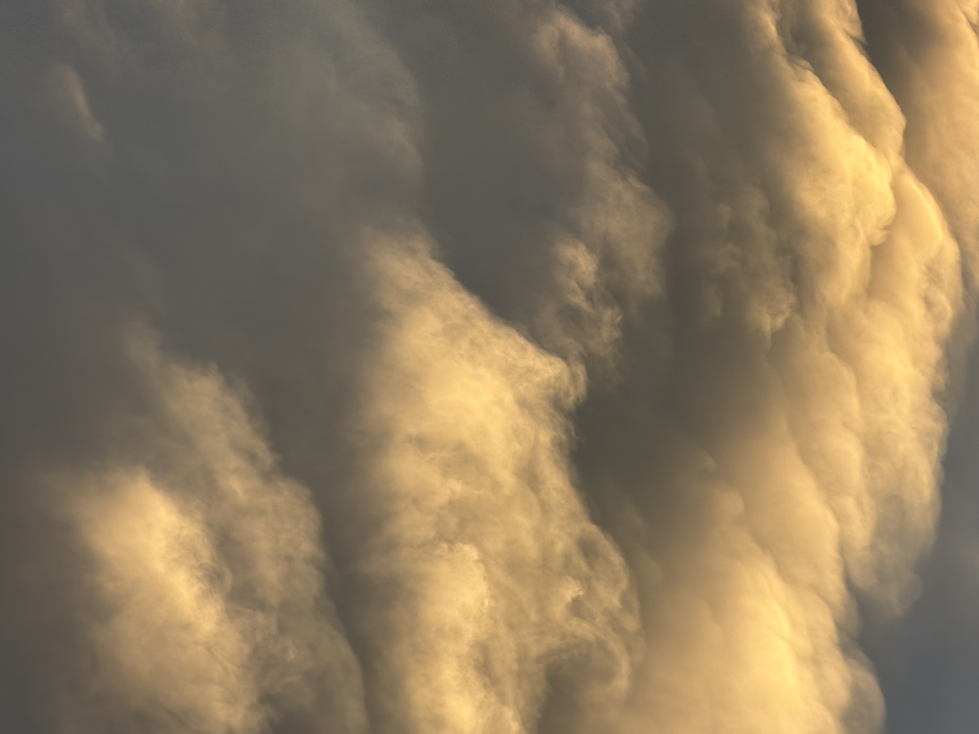 The image shows a dramatic sky filled with turbulent clouds, illuminated by a warm, golden light, likely from a sunset or sunrise. The clouds are textured and appear to be rolling or rippling, with darker, gray clouds forming a stark contrast to the brightly lit areas. The overall impression is one of atmospheric intensity. The scene is entirely composed of the sky; there are no discernible earthly elements like buildings, landscapes, or people. The location is specified as Çolpon-Ata, Kyrgyzstan, but this information is contextual and not visually present in the photograph.