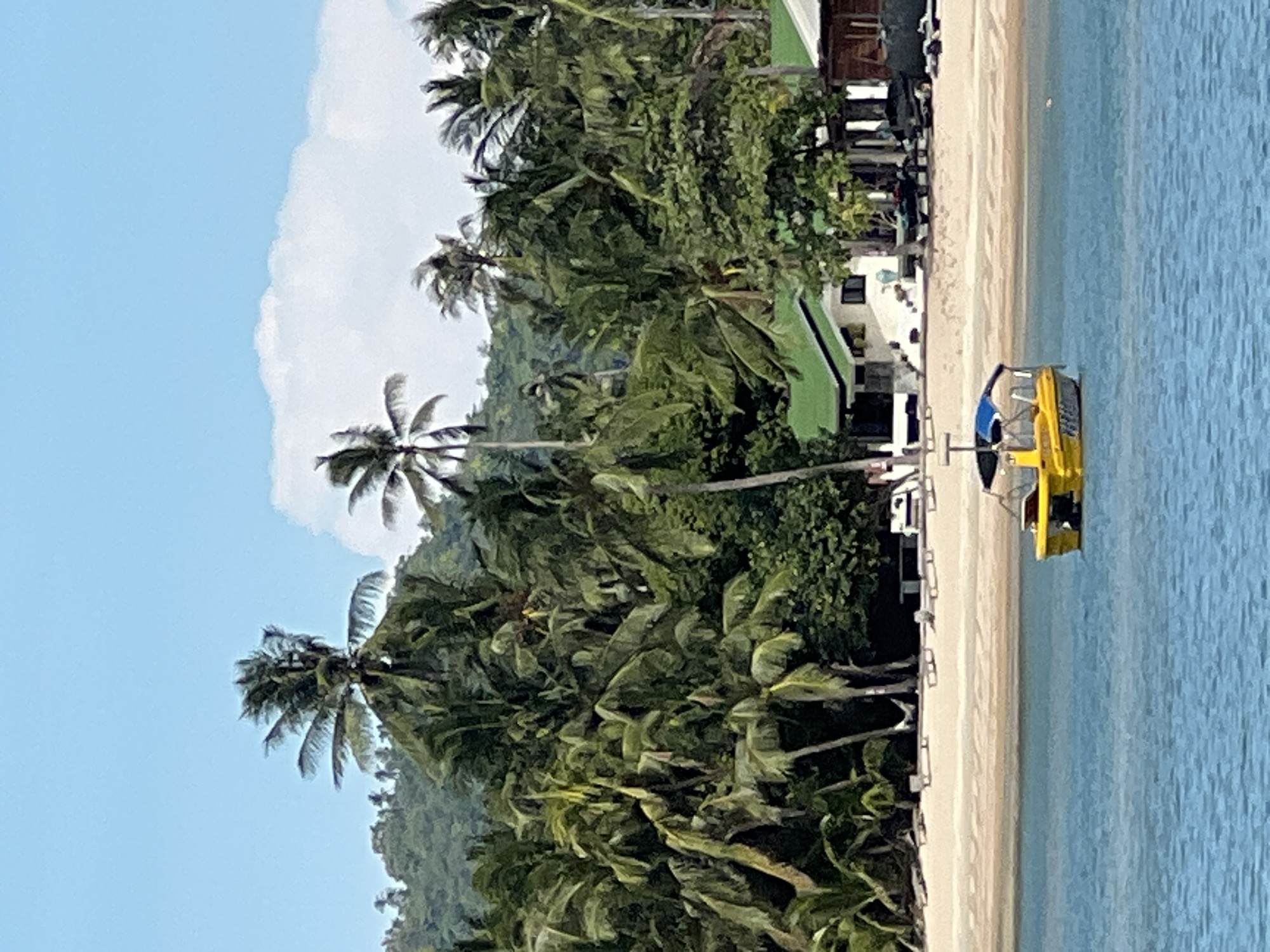 This image depicts a serene tropical scene at Baan Hua Thanon, Thailand. In the foreground, a bright yellow boat with a blue canopy floats on calm, blue water. The boat has visible lettering on its side, which is difficult to decipher due to image quality. The middle ground features a sandy beach lined with lounge chairs and umbrellas, suggesting a resort or recreational area. Behind the beach, a dense jungle of palm trees and other lush vegetation covers a rolling hillside, creating a verdant backdrop. Several buildings with green roofs are visible amidst the trees, indicative of the local architecture and possibly tourist accommodations. The sky is a clear blue, with a few white clouds scattered across it. The overall atmosphere is peaceful and idyllic, characteristic of a tropical island paradise.