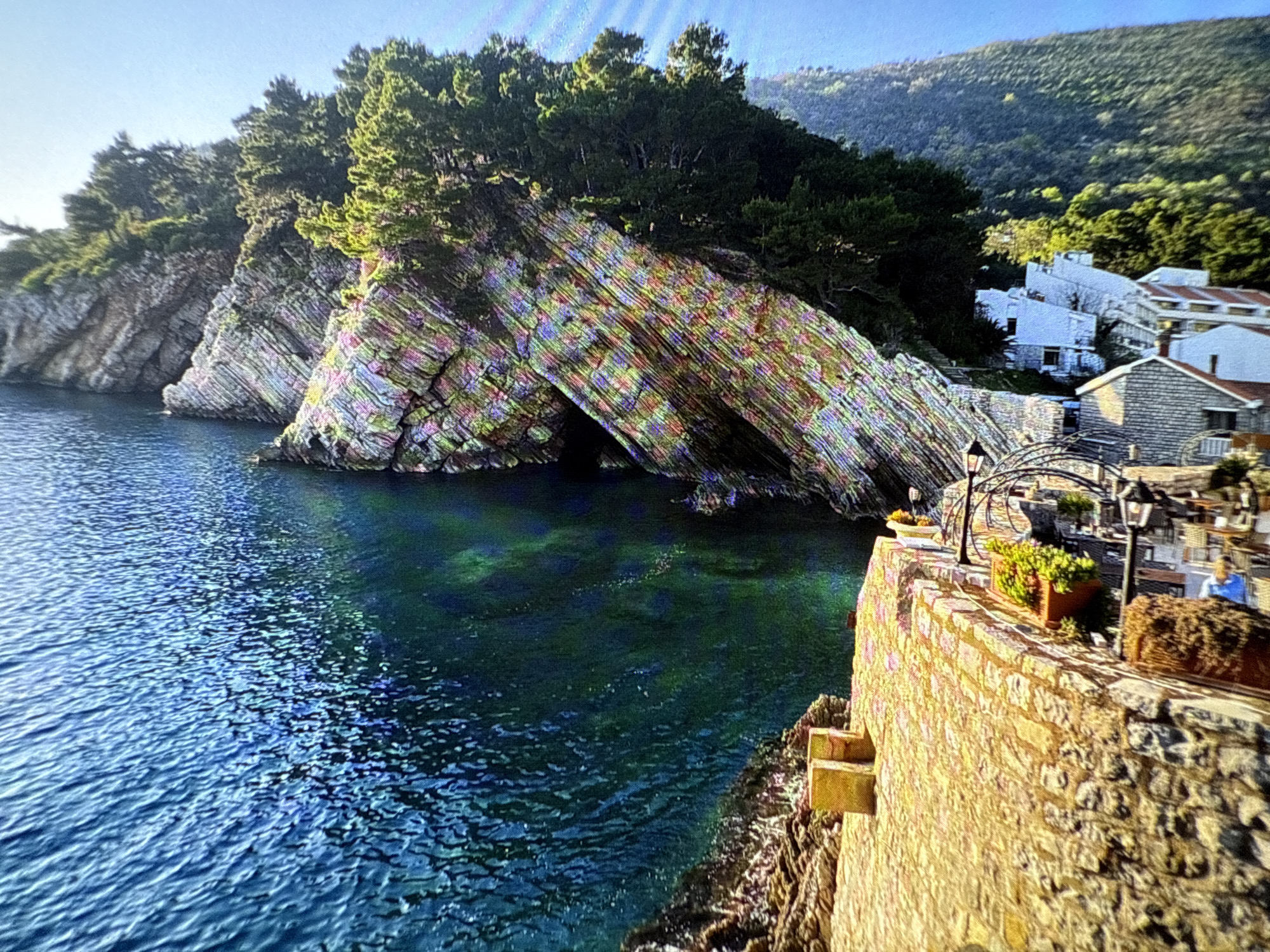 The image captures a scenic view in Budva, Montenegro. On the left, there is a large, layered rock formation with a small cave at its base, topped with evergreen trees. The rock face displays distinct horizontal strata in shades of beige, grey, and brown. The blue-green water of the Adriatic Sea laps against the rocks. To the right, a stone wall runs along the edge of the scene, with a black lamppost and planters with greenery. In the background, whitewashed buildings with red-tiled roofs are partially visible, nestled against a hillside covered in green vegetation. The overall impression is one of a coastal landscape with a mix of natural and built environments.