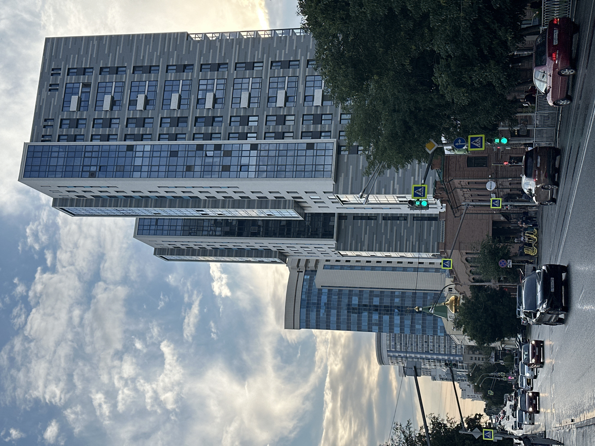 The image depicts a city street in Yekaterinburg, Russia, characterized by a mix of modern high-rise buildings and older architecture. The street is wet, suggesting recent rain, and traffic is present with several cars driving in both directions. Pedestrians are visible on the sidewalks. Traffic lights and pedestrian crossing signs are prominent features of the scene. In the background, a golden-domed building, likely a church, stands out amidst the urban landscape. The sky is filled with dramatic clouds, casting a moody atmosphere over the city.
