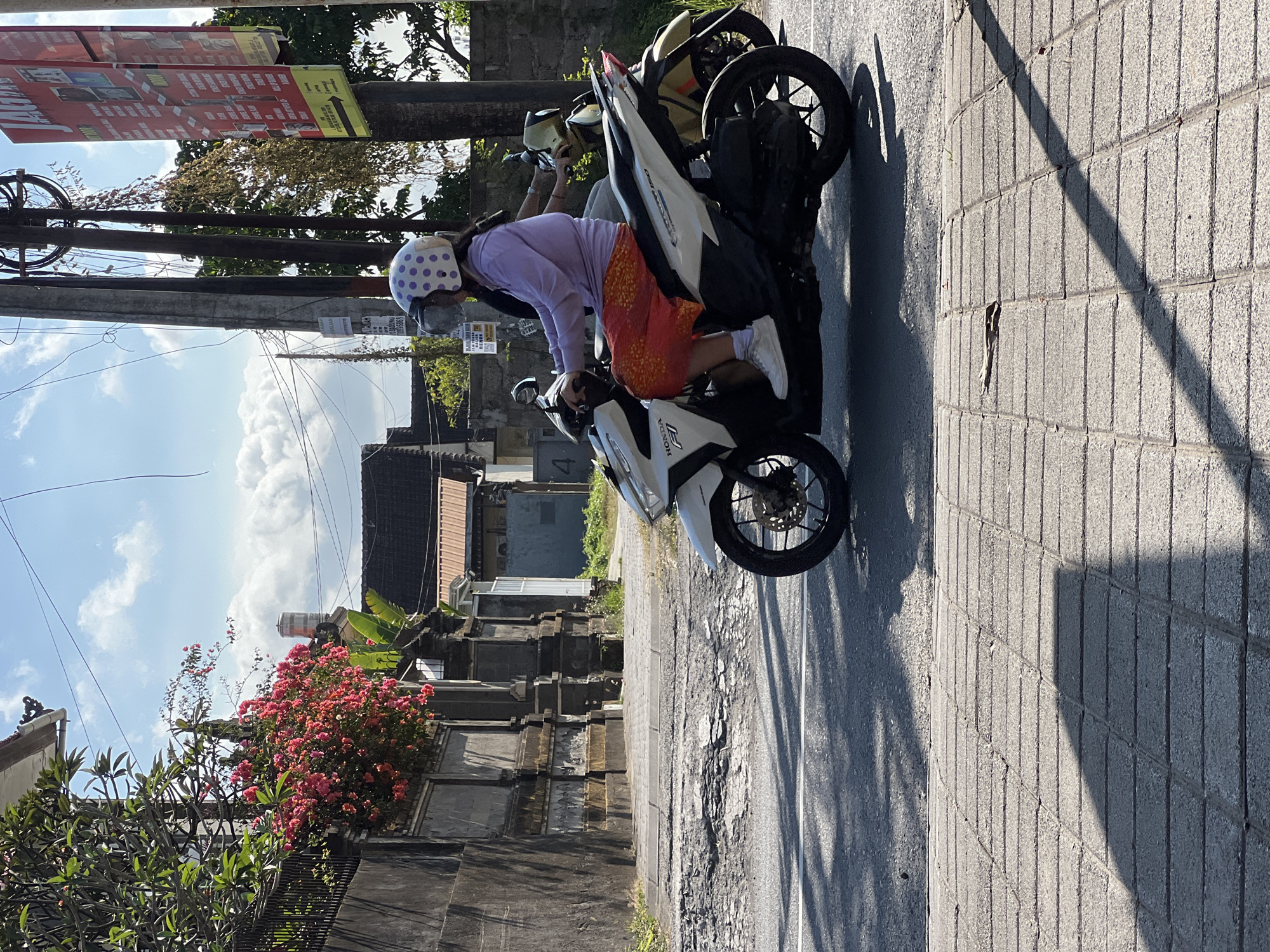 A person wearing a white helmet with purple polka dots rides a white Honda scooter on a paved road in Denpasar, Indonesia. The rider is wearing a light purple long-sleeved shirt and an orange skirt with a patterned design. They are also wearing white sneakers. The scooter has the "HONDA" logo visible on the side. In the background, there is a stone wall with ornate carvings, a cluster of pink flowers, and trees. Several utility poles with numerous wires are also visible. The road has a cracked asphalt surface, and the sidewalk is made of grey bricks. Sunlight casts shadows from trees and the utility poles onto the road and sidewalk. Another scooter, mostly yellow, is visible behind the main scooter, ridden by a second person. The sky is blue with fluffy white clouds. A red and white advertisement banner is attached to one of the utility poles.