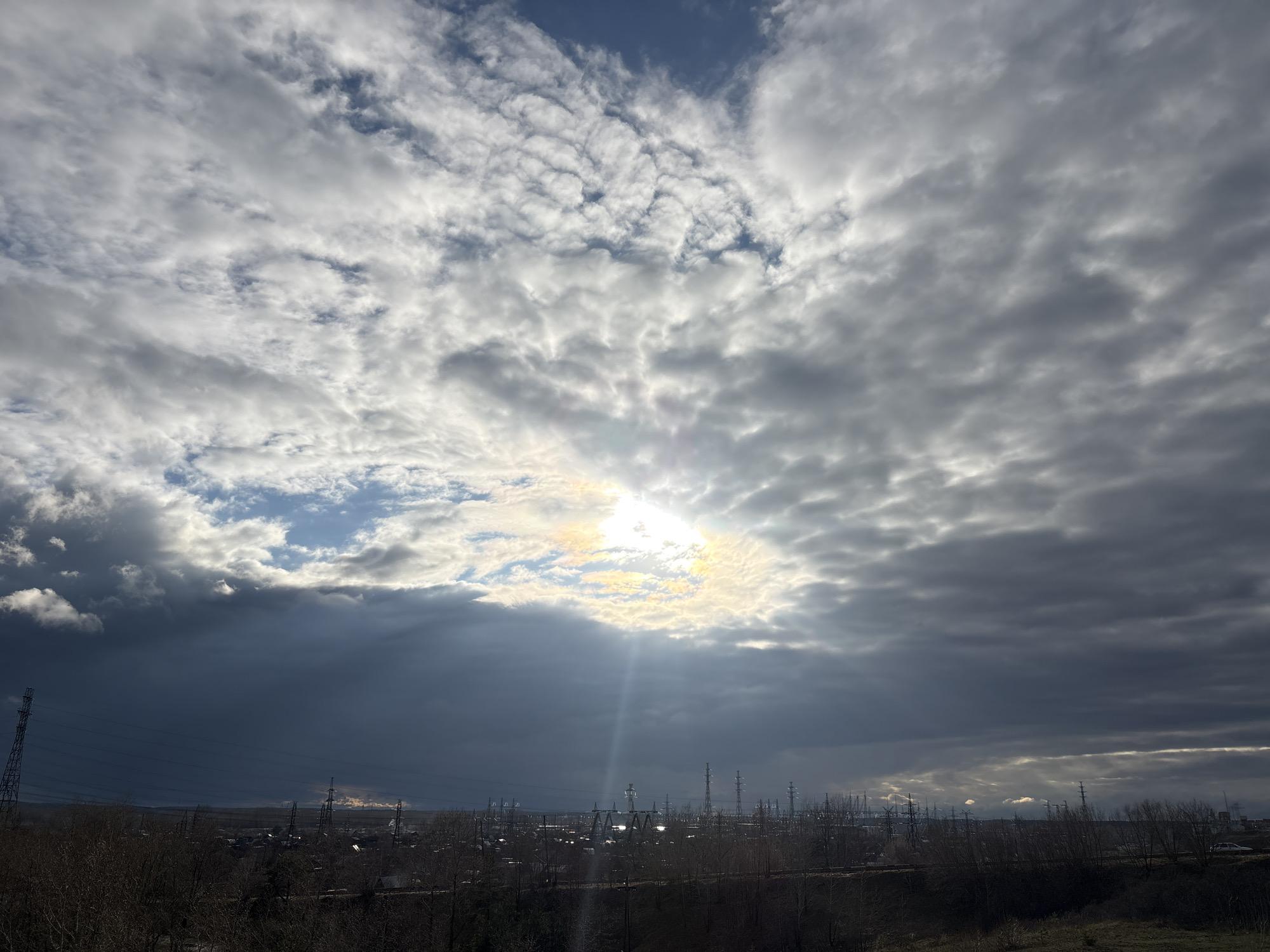 The image displays a dramatic and expansive outdoor scene dominated by a dynamic sky over an industrial landscape. The sky is filled with a dense cover of clouds, ranging from bright white and fluffy formations to darker, heavier grey masses, with patches of clear blue sky visible in the upper sections. A central, intense burst of sunlight breaks through a prominent opening in the clouds, casting distinct rays downwards towards the horizon.
Below the horizon, an extensive landscape unfolds, featuring numerous tall electrical pylons and power line towers of various designs, stretching across the mid-ground. In the deeper distance, industrial structures and buildings are discernible, with what appears to be smoke or steam rising from multiple points, particularly towards the right side of the frame where sunlight catches some emissions. The immediate foreground and mid-ground consist of bare trees and sparse vegetation, suggesting a late autumn or winter season. The ground cover is dry and earthy.
The scene depicts a moment of dramatic weather, likely daytime, where the sun is momentarily piercing through heavy cloud cover, creating a striking contrast between light and shadow. No discernible human activity or visible text is present. The setting appears to be an industrial or semi-urbanized area, consistent with the provided location context of Chaikovsky, Russia.