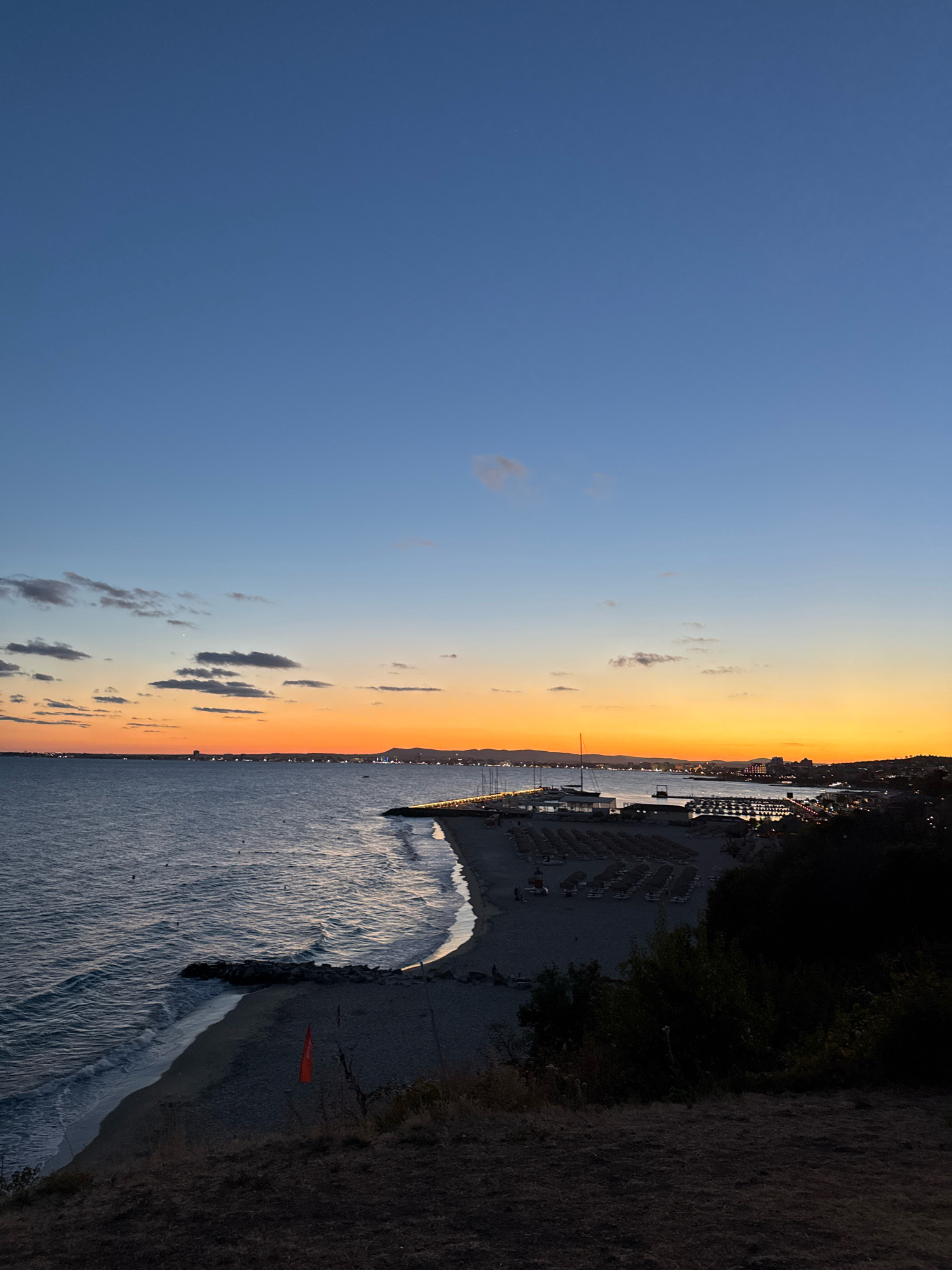 The image captures a scenic view of the beach and coastline in Sveti Vlas, Bulgaria, during dusk. The sky is a gradient of deep blue at the top, transitioning to vibrant orange and yellow hues near the horizon, indicative of a sunset. Scattered clouds are visible in the upper part of the sky and also along the horizon.
The sea is calm with gentle waves lapping the shore. The beach itself is a mix of sand and pebbles, with rows of empty sun loungers and umbrellas neatly arranged, suggesting it was a popular spot earlier in the day. A small red flag is planted on the beach in the foreground.
In the distance, a town or city is visible along the coastline, illuminated by lights. A pier extends into the sea, with several sailboats and yachts docked nearby. The landscape on the right side of the image is darker, with trees and vegetation obscuring the view further inland. The overall atmosphere is tranquil and picturesque, with the fading light casting a warm glow over the scene.