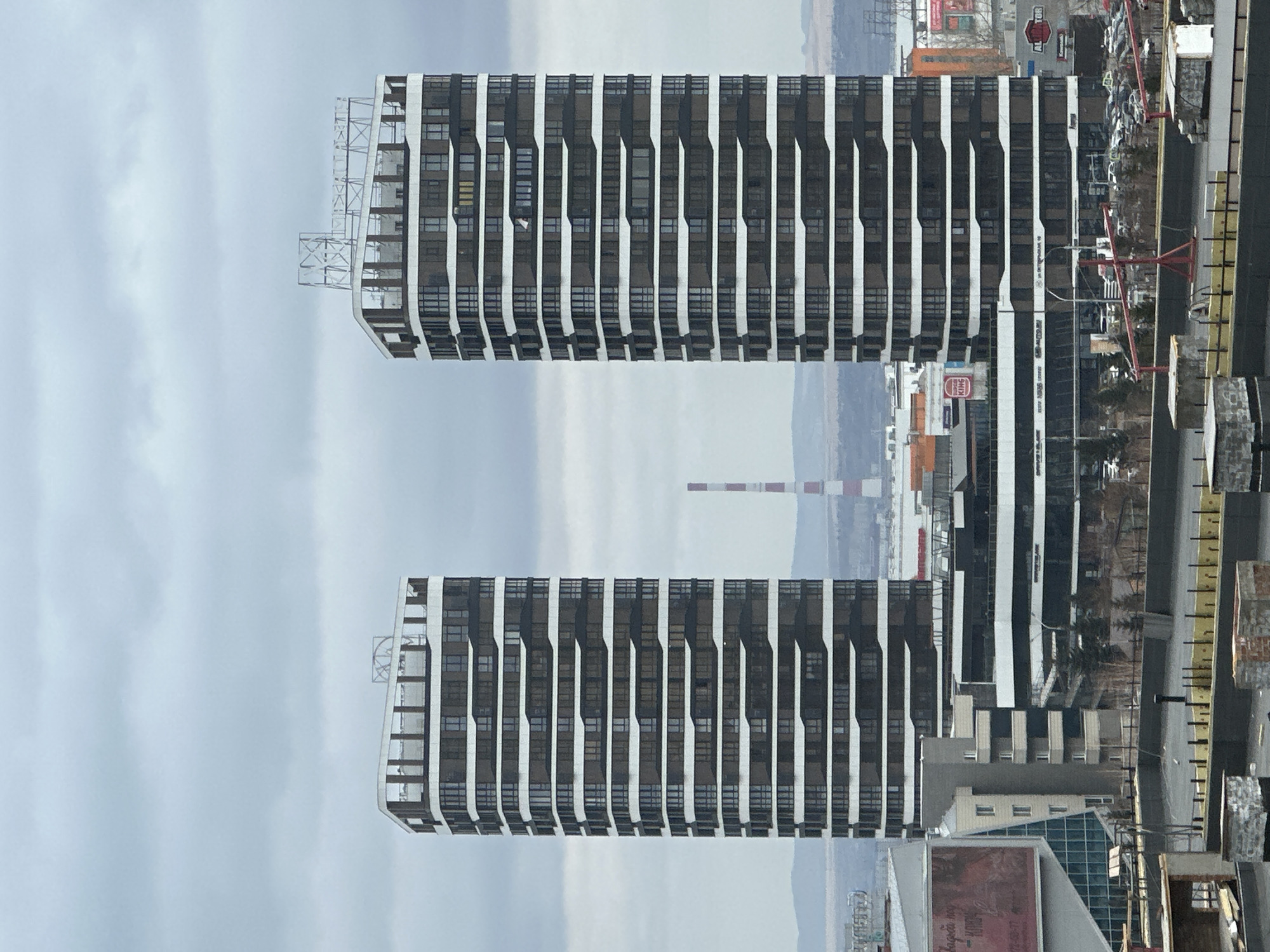 The image depicts a view of buildings in Krasnoyarsk, Russia. Two nearly identical, modern high-rise buildings with a distinctive black and white striped facade dominate the scene. Each building appears to have a curved edge on one side, possibly for balconies. A metal structure is visible on the top edge of one of the buildings.
In the background, there are other buildings, including industrial structures like a smokestack with red and white stripes. The sky is overcast, suggesting a cloudy day. There's evidence of urban life on the ground including infrastructure, commercial signs (including a King Burger), and buildings of varying sizes and architectural styles. The image seems to be taken from an elevated vantage point.