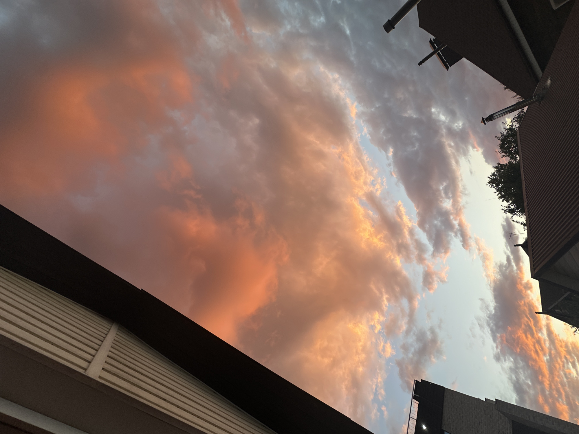 This media file captures a dramatic sky at dusk, with the clouds painted in vibrant hues of orange, pink, and gray. The sun appears to be setting, casting a warm glow on the clouds and creating a sense of awe.
The scene is framed by the dark silhouettes of buildings, their sharp lines contrasting with the soft, amorphous shapes of the clouds. The architecture suggests an urban environment, with elements like corrugated siding and what appear to be ventilation pipes or chimney flues visible on the roofs. The perspective is looking upwards, emphasizing the grandeur of the sky.
The time of day is clearly twilight, as indicated by the rich colors in the sky and the subdued light. The weather appears to be partly cloudy, with the clouds actively participating in the spectacle of the sunset. There are no people or specific activities depicted; the focus is solely on the natural phenomenon of the sky.
The emotional tone evoked is one of tranquility and wonder. The vibrant colors and the vastness of the sky create a sense of peace and contemplation. The image captures a fleeting moment of natural beauty, a common experience during sunset.
The location context is Nizhny Novgorod, Russia, though this is not visually apparent from the image itself. There is no visible text within the media file.