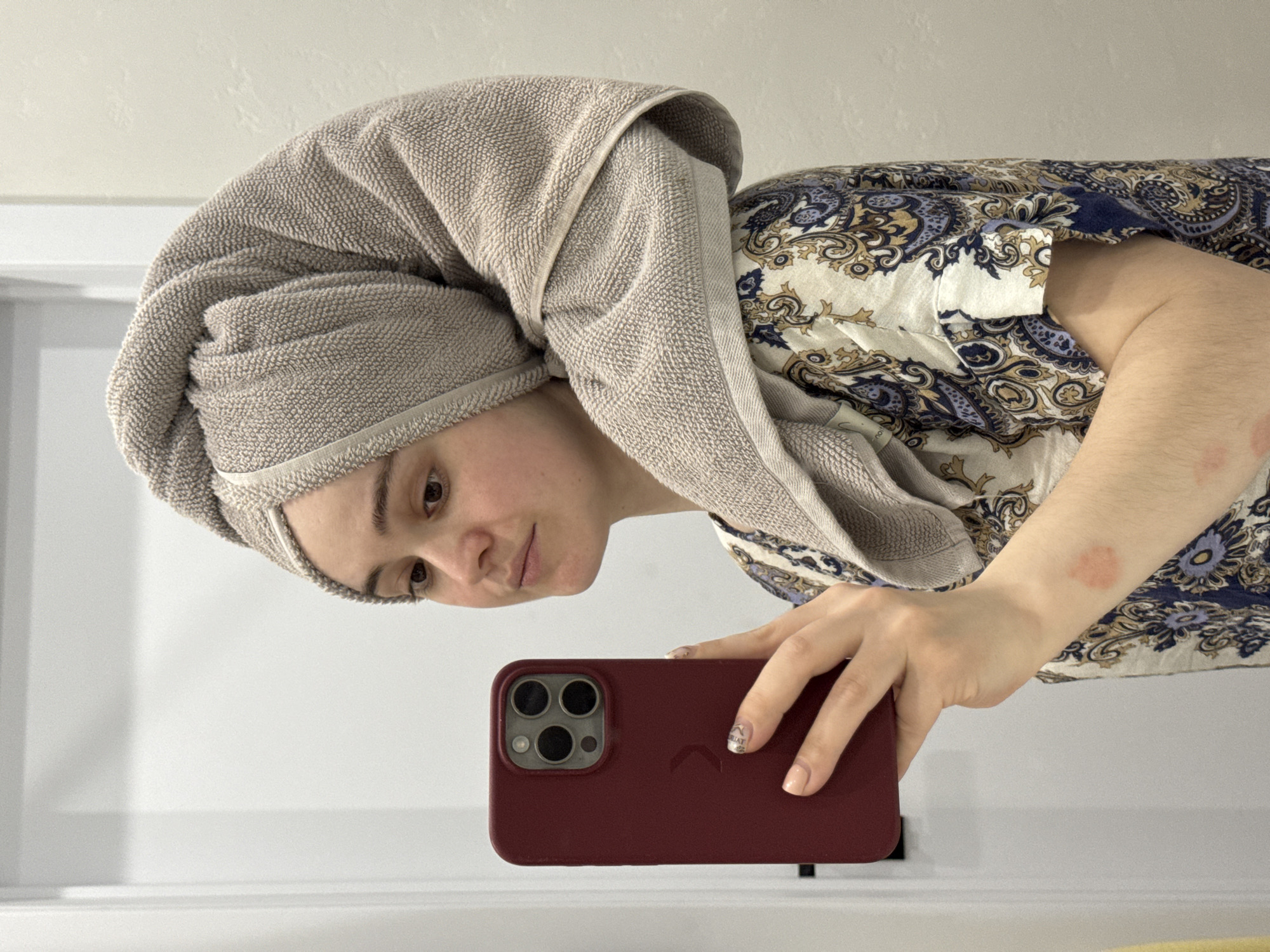 A person is taking a selfie. The person has a beige towel wrapped around their head and is wearing a patterned shirt. They are holding a burgundy-colored smartphone in their right hand. The background appears to be a white wall, possibly in a bathroom or similar space. There is no indication of the city of Izhevsk, Russia, in this image.