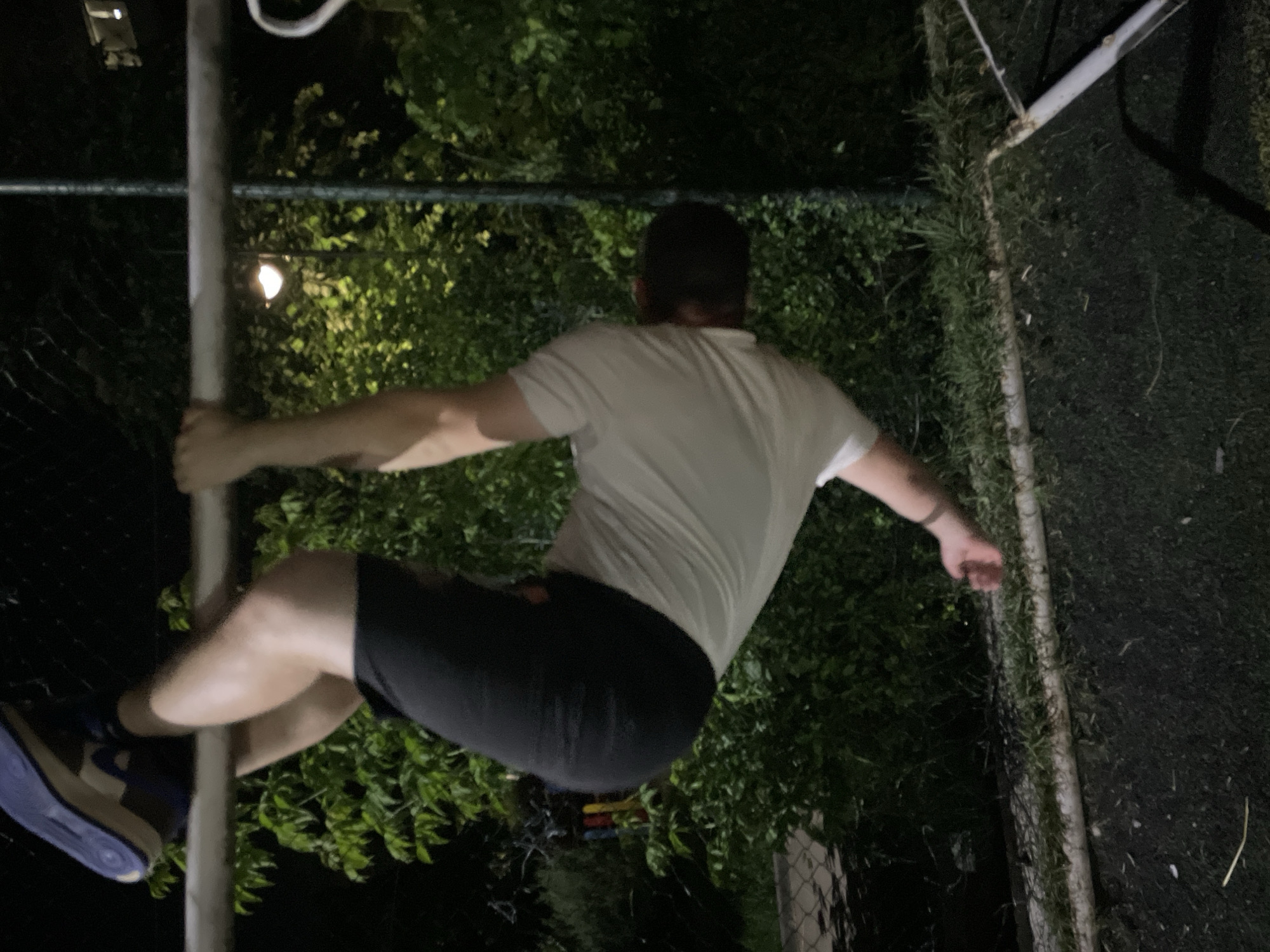 The image displays an outdoor scene at night in Razgrad, Bulgaria. A male individual is depicted hanging upside down from a horizontal, silver-colored metal bar. He is wearing a white short-sleeved t-shirt, dark shorts, and blue athletic shoes. His legs are hooked over the top of the bar, and his hands are gripping it for support. The individual's head is pointed downwards, away from the camera. The background is dominated by dense green foliage, suggesting trees and bushes, dimly illuminated by an artificial light source, specifically a street lamp, visible behind the bar. The ground surface directly beneath the bar is dark, possibly asphalt or a rubberized court, with a horizontal strip of green grass running along its edge. A light-colored, pipe-like structure rests on the ground to the right of the grass strip. A chain-link fence is partially visible on the far left side of the frame.