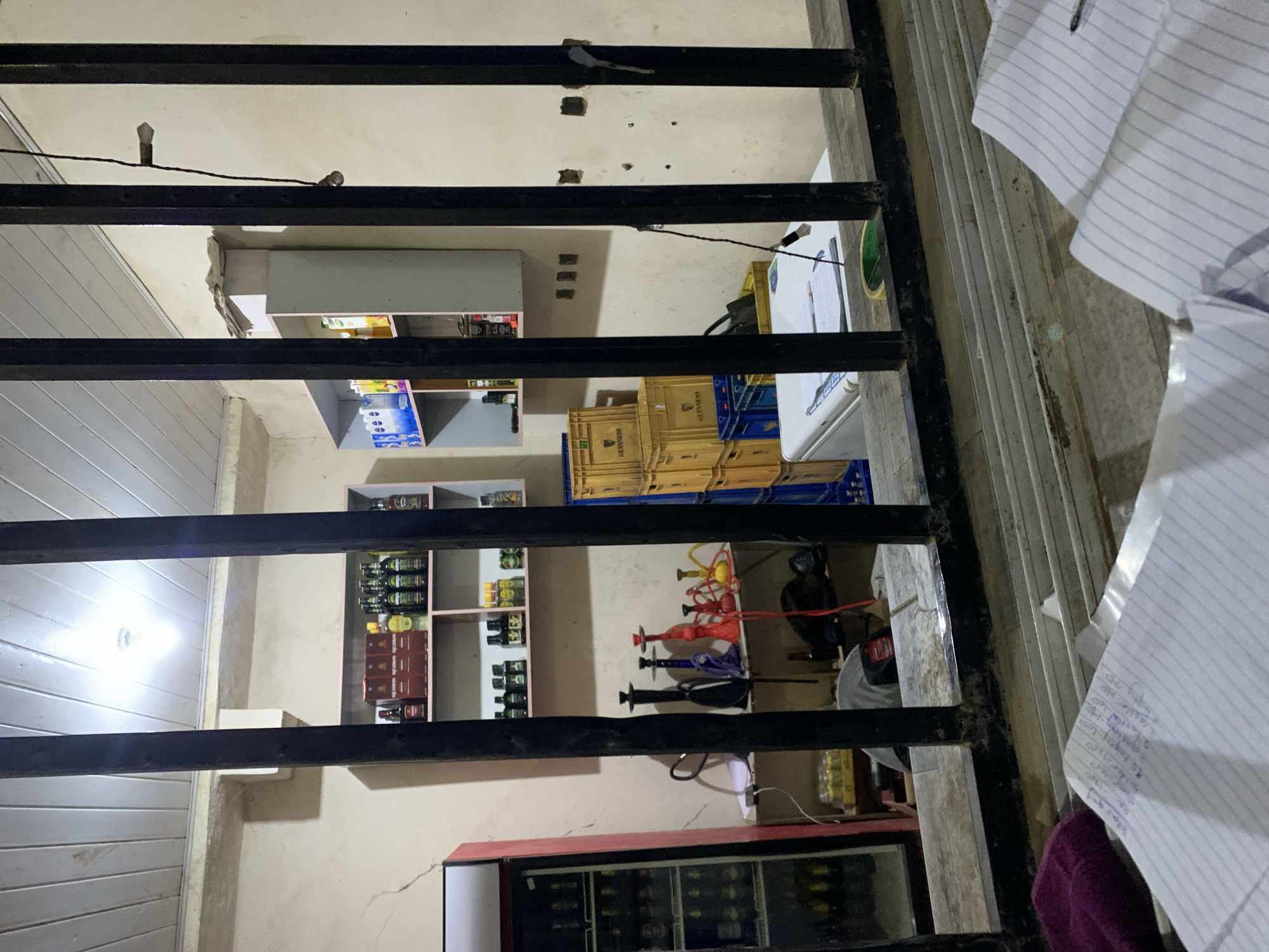 The image depicts the interior of a small retail establishment in Kabong, Nigeria, viewed from behind a wooden counter or window sill with three prominent black vertical metal bars.
In the immediate foreground, a worn wooden surface, possibly a counter, is present. On its left, a purple/maroon fabric item rests, while on the right, several sheets of lined paper with handwritten notes and a pen are visible.
Beyond the bars, the shop's interior features light-colored, somewhat worn walls with visible cracks and imperfections. The ceiling is constructed from horizontal wooden planks, with a single bright, bare light bulb providing illumination.
To the left, a tall, red-framed commercial display refrigerator with a glass door is stocked with numerous bottled beverages. Towards the center-left, a shelf holds an array of colorful shisha pipes (hookahs) in black, blue, red, and yellow. Below this, a red cord is visible.
On the back wall, two shelving units are present: a pale pink unit on the left displaying various bottled alcoholic beverages and other products, and a light grey unit on the right holding bottles with blue (possibly Nivea) and yellow labels (possibly juices or oils).
To the right, a stack of yellow plastic crates, some bearing "GUINNESS" branding, is visible, along with blue plastic crates. A white chest freezer or cooler is situated below these crates, with papers and a small object on its surface. A dark cord with a small cylindrical attachment hangs from the ceiling or wall on the right side, which also exhibits several square indentations and marks.
The back of a person, wearing a grey shirt, is partially visible in the lower mid-ground behind the counter/bars, suggesting someone is present or working within the shop. The lighting is artificial, originating primarily from the overhead bulb.