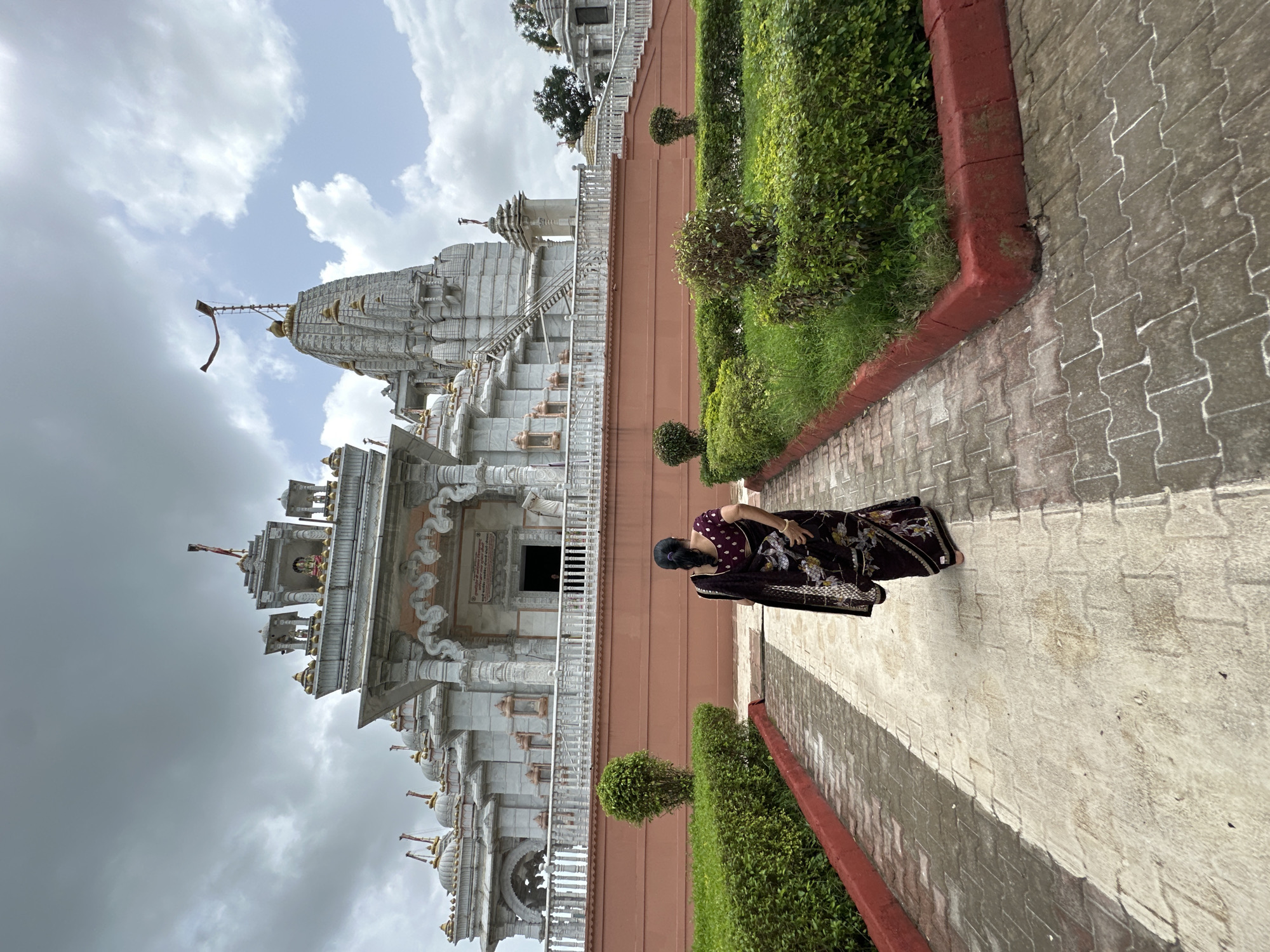 The image depicts the exterior of a Hindu temple in Shahapur, India. In the foreground, a woman wearing a dark patterned sari walks away from the camera on a paved pathway. She is positioned on the left side of the path, with well-maintained green bushes and hedges flanking both sides. The pathway is constructed with interlocked concrete pavers.
The temple itself is a large, ornate structure made of light-colored stone, possibly marble. It features multiple tiered roofs, decorative spires, and intricate carvings. A tall central tower, also adorned with smaller spires, dominates the skyline of the temple complex. A red flag is visible at the very top of the main tower. Steps lead up to the entrance of the temple, and a railing runs along the elevated platform.
The sky above is overcast with a mix of dark and lighter clouds. The overall scene suggests a religious site within an urban or semi-urban environment, indicated by the presence of manicured landscaping surrounding the temple. The lighting suggests it is daytime, possibly late morning or afternoon, given the cloud cover.