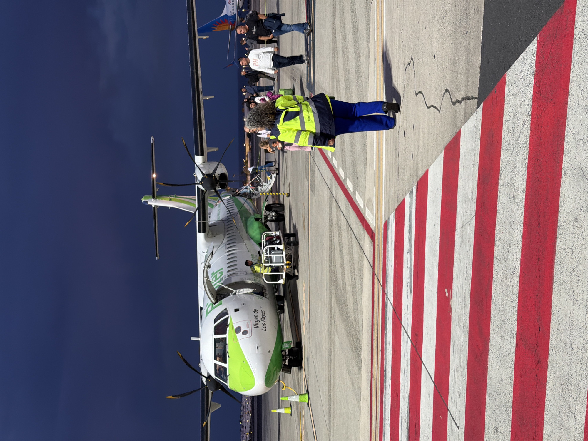 The image captures an airport scene, likely during late afternoon or early evening, featuring a white and green turboprop aircraft on the tarmac. The aircraft, which has propellers, is labeled "Virgen de Los Reyes" on its fuselage in green lettering.
In the foreground, a ground crew member, identifiable by a bright yellow high-visibility vest with partial text "Aeropuerto de..." visible on the back, stands with their back to the viewer. They wear blue trousers and black shoes. Directly behind this individual and around the aircraft's open door, several people, presumably passengers, are visible, suggesting boarding or disembarking activity. Another aircraft can be seen partially in the background, further indicating an airport setting.
The tarmac is marked with distinct red and white striped lines, which are common safety or boundary markings at airports, along with other yellow lines and a couple of small safety cones near the aircraft's nose gear. The sky overhead is a deep, clear blue, suggesting good weather conditions. The overall scene depicts a typical operation on an airport ramp.