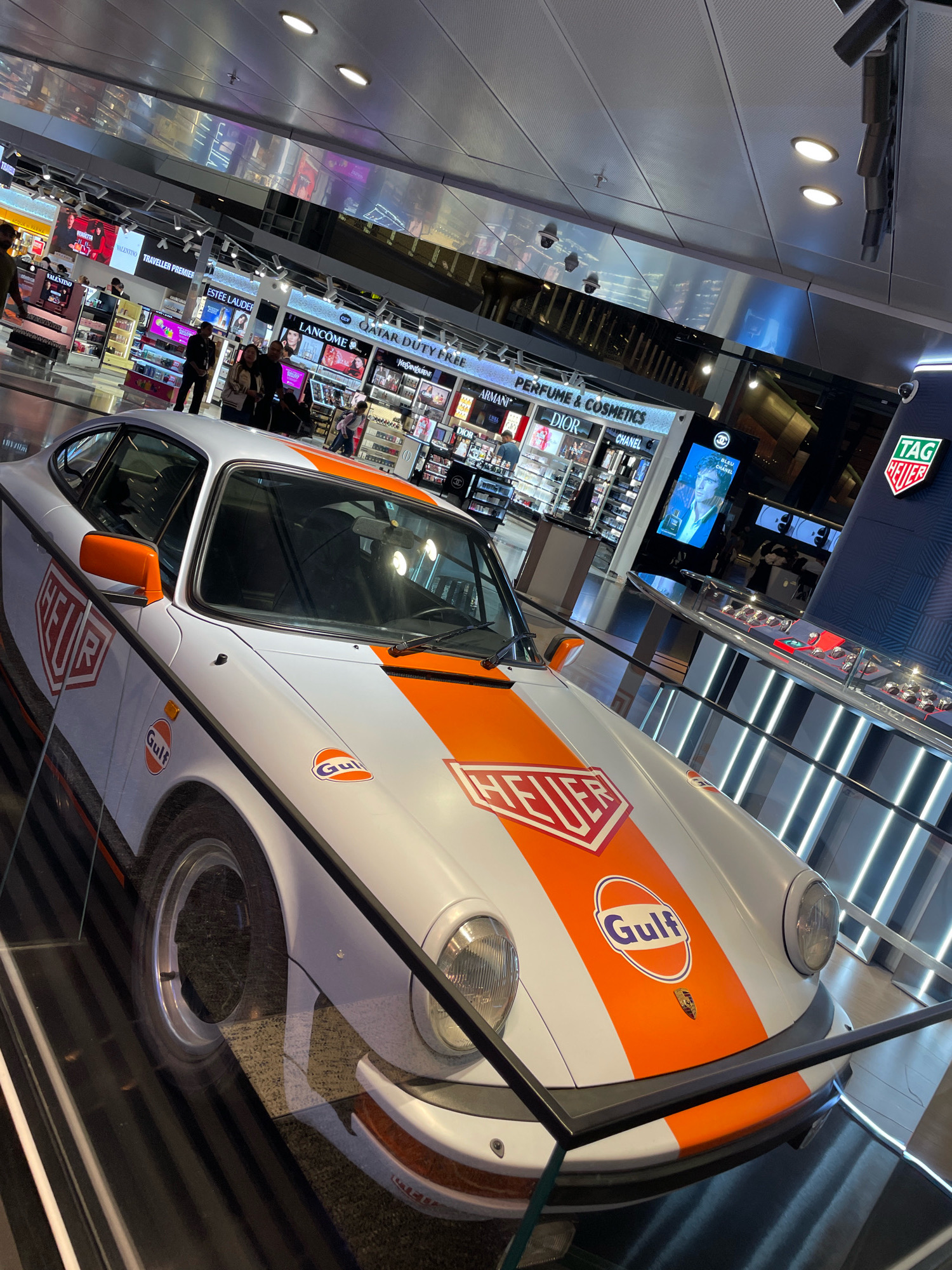 The image shows a vintage Porsche 911 race car, painted white with prominent orange stripes and Gulf Oil livery, displayed indoors. The car appears to be a centerpiece within what seems to be a luxury retail environment, possibly an airport duty-free shop. In the background, shelves stocked with various perfumes and cosmetics are visible, along with branding for "Qatar Duty Free," "Estée Lauder," "Lancôme," "Dior," and "Chanel." A prominent "TAG Heuer" advertisement is also present. Several individuals are seen browsing or walking through the retail space. The car is sectioned off by a black metal railing and glass barrier, suggesting it is an exhibit. The overall lighting is bright and modern, consistent with a high-end commercial setting. The presence of "Qatar Duty Free" strongly indicates that the location is Hamad International Airport in Doha, Qatar.