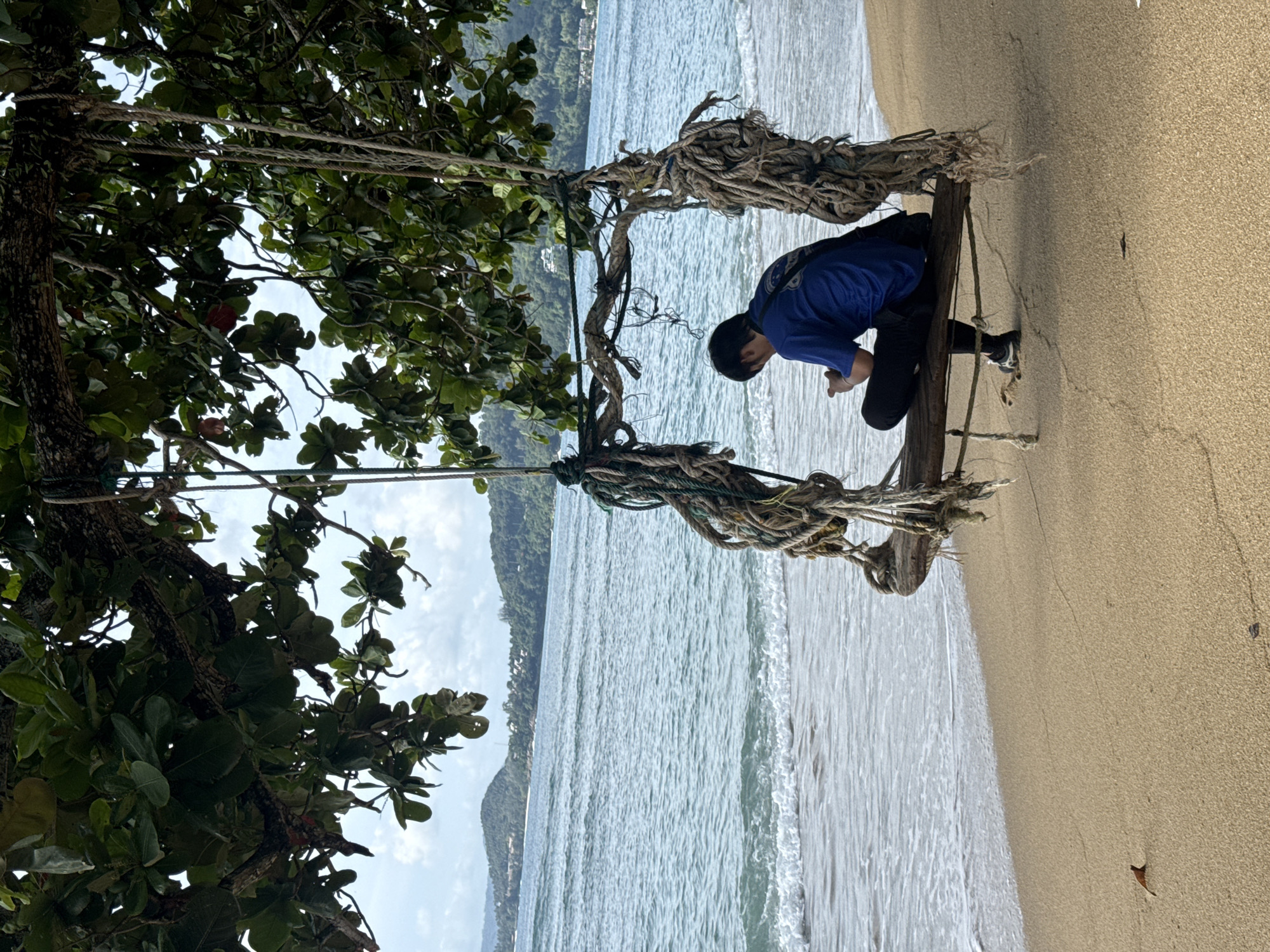 A person sits on a wooden swing attached to a tree on a sandy beach in Kamala, Thailand. The person is wearing a blue t-shirt and black pants and is looking down at their phone. The swing is made of weathered wood and thick ropes, suspended from the branches of a large tree with green leaves. The beach has fine, light-brown sand, and the ocean is a calm turquoise with gentle waves lapping at the shore. In the background, lush green hills rise, dotted with buildings, under a cloudy sky. The scene captures a moment of quiet contemplation by the sea.