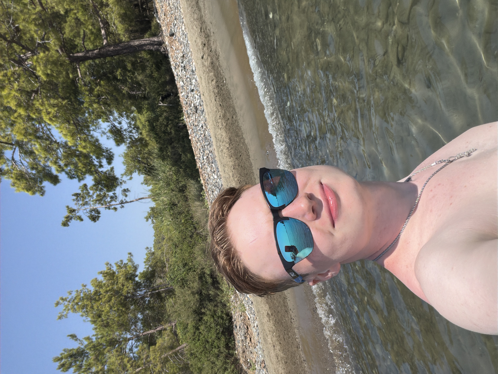 A young person with fair skin and reddish-blonde hair is taking a selfie while standing in clear, shallow water. They are wearing blue-tinted sunglasses with black frames, which reflect the surroundings. A silver chain with a cross pendant is visible around their neck. The person's upper body shows signs of sunburn, particularly on their shoulder and neck. The background consists of a pebbly beach with sandy patches, lush green trees, and a clear blue sky. The scene suggests a sunny day at a coastal location, likely a beach in Kemer, Turkey, given the vegetation and clear water typical of the region.