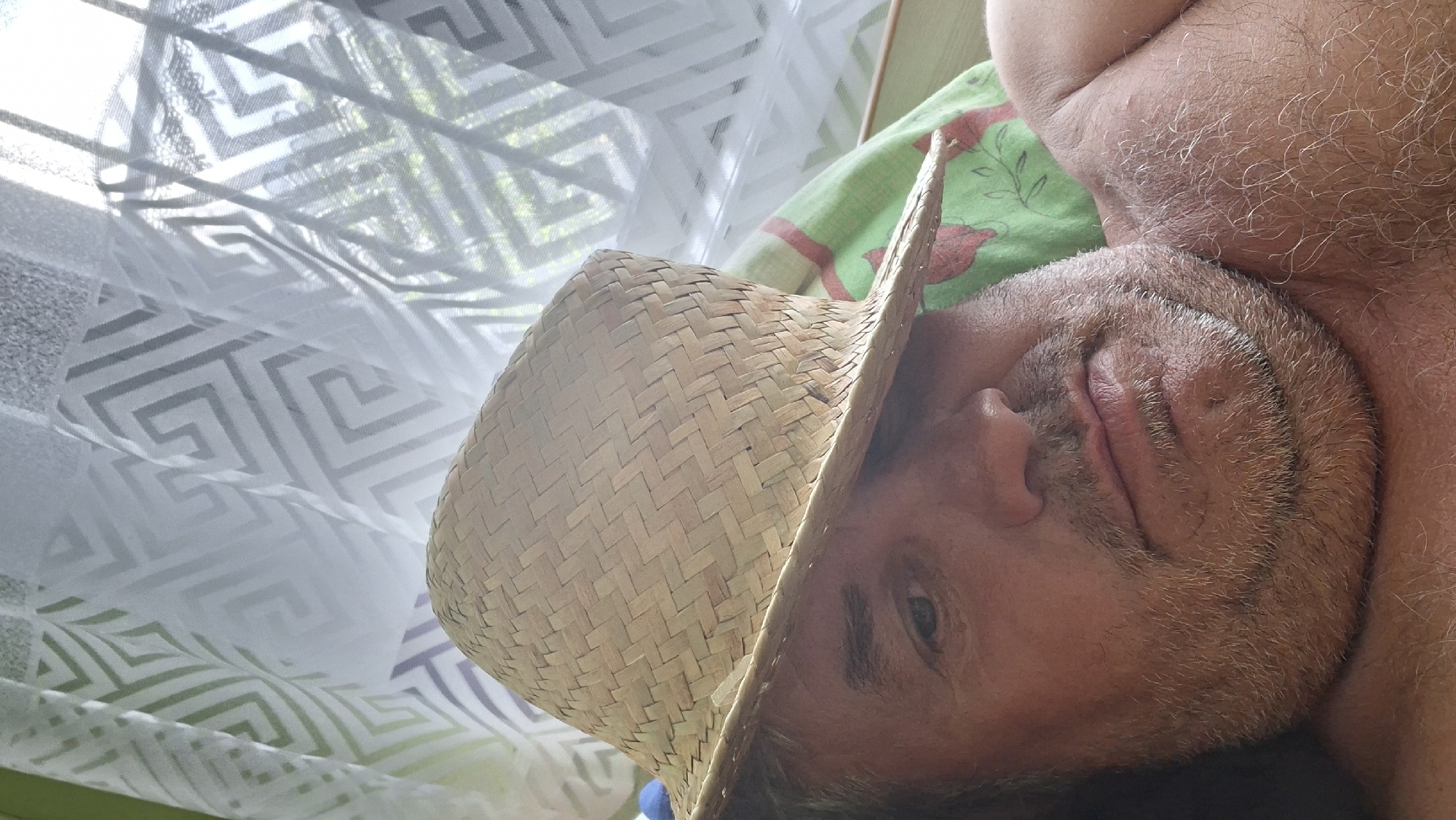 The image is a close-up, selfie-style photograph of a man wearing a straw hat that partially obscures his face. The man appears to be outdoors, possibly lying down, with a patterned curtain or sheer fabric in the background, which is translucent and shows light and some greenery filtering through. The man has graying stubble on his face and chin. He is shirtless, and his skin appears tanned. The overall lighting suggests it is daytime. There are no visible landmarks or contextual clues within the image that would indicate the location in the city of Voinova, Moldova.