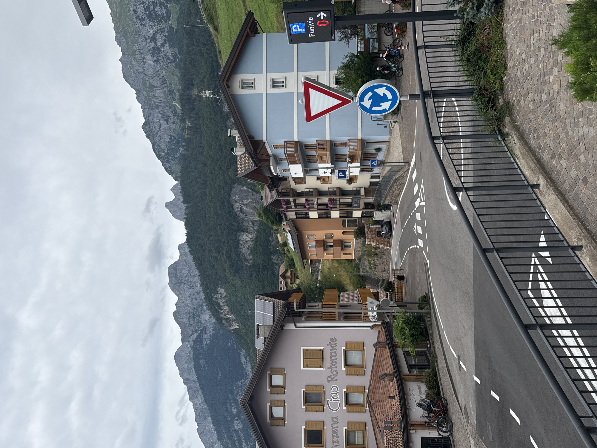 The image depicts a street scene in the city of Molveno, Italy, set against a backdrop of imposing mountains. In the foreground, a dark metal railing overlooks a cobblestone pathway and a road with clear white lane markings. The road curves to the right, leading towards a pedestrian crossing marked with white arrows.
Several buildings line the street. On the left, a light purple building features windows with wooden shutters and a sign that reads "Pizzeria Ciao Ristorante". Adjacent to it, a building with a red-tiled roof also has windows and shutters. To the right, a light blue building with white trim has multiple floors, balconies, and windows. Further down the street, an orange building is visible.
Traffic signs are prominent: a large red and white yield sign is mounted on a pole, accompanied by a blue circular sign with three arrows indicating a roundabout. A blue sign with a white 'P' and the word "Funivie" (cable car) along with a red arrow pointing to the right is also visible, indicating directions.
On the road, several bicycles are parked. In the distance, a few people are visible, including two individuals on bicycles near the traffic signs. The sky is overcast, and the mountains in the background are covered in lush green trees up to a certain elevation, with rocky peaks rising above them. The overall atmosphere suggests a quiet, possibly tourist-oriented town in a mountainous region.