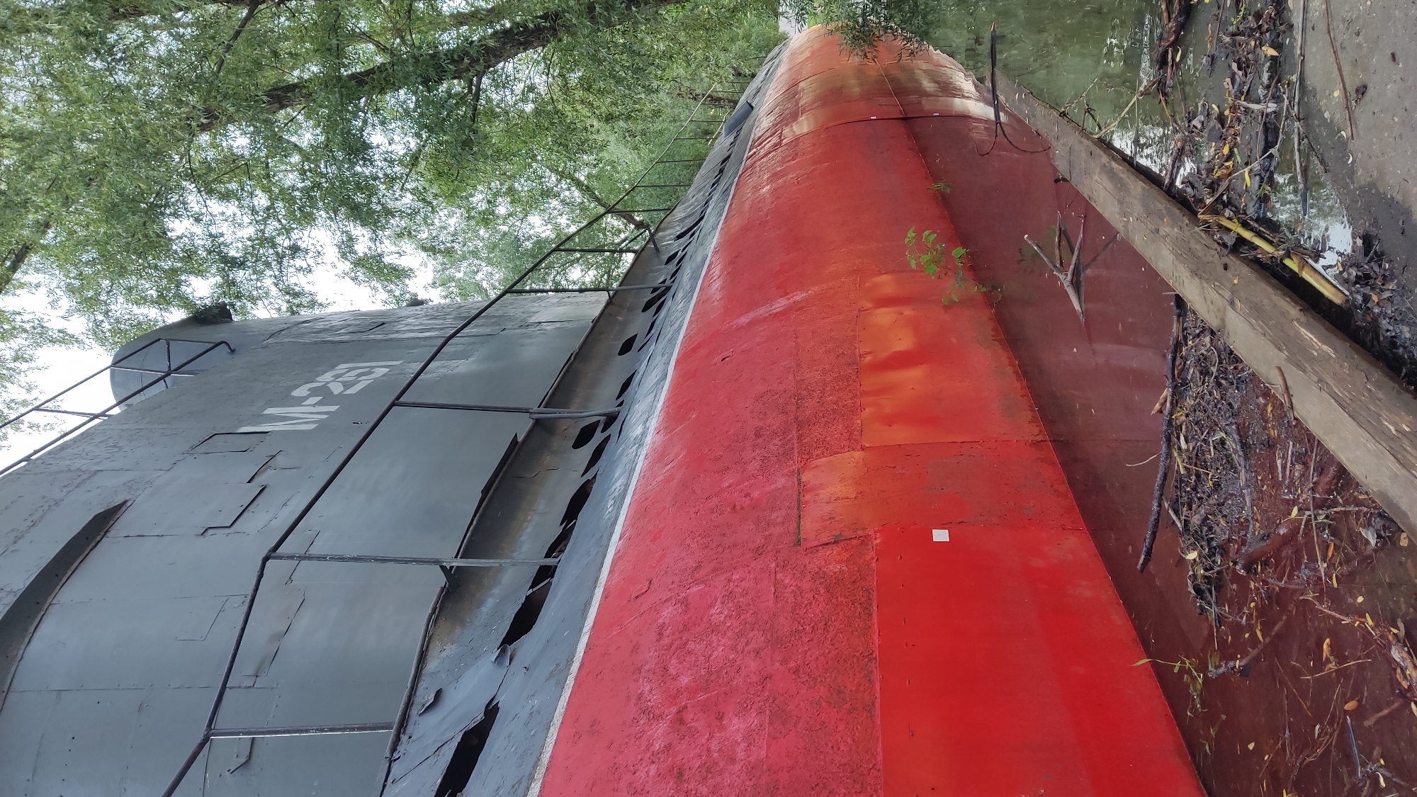 The image shows the submerged submarine M-261 in Krasnodar, Russia. The submarine is lying on its side, partially submerged in water filled with debris. The hull is painted in a dark gray and red color scheme. The inscription "M-261" is visible on the hull. Trees and vegetation surround the submarine, partially obscuring the view. A wooden beam lies along the side of the submarine in the water.