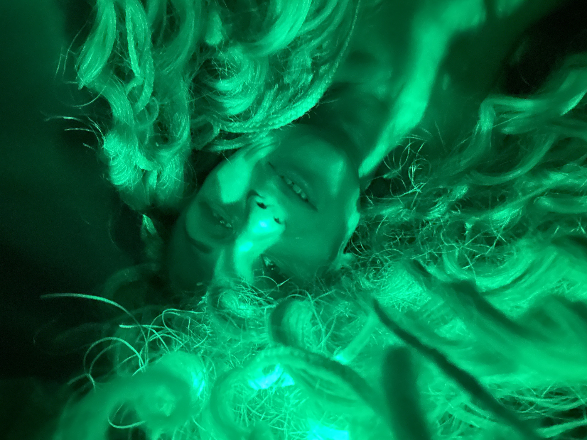 A person lies on their back amidst a tangled mass of what appears to be hair, with a green cast across the entire image. The hair seems voluminous and unkempt, obscuring much of the background. The person is smiling and facing the camera. There is no identifiable indication of location.