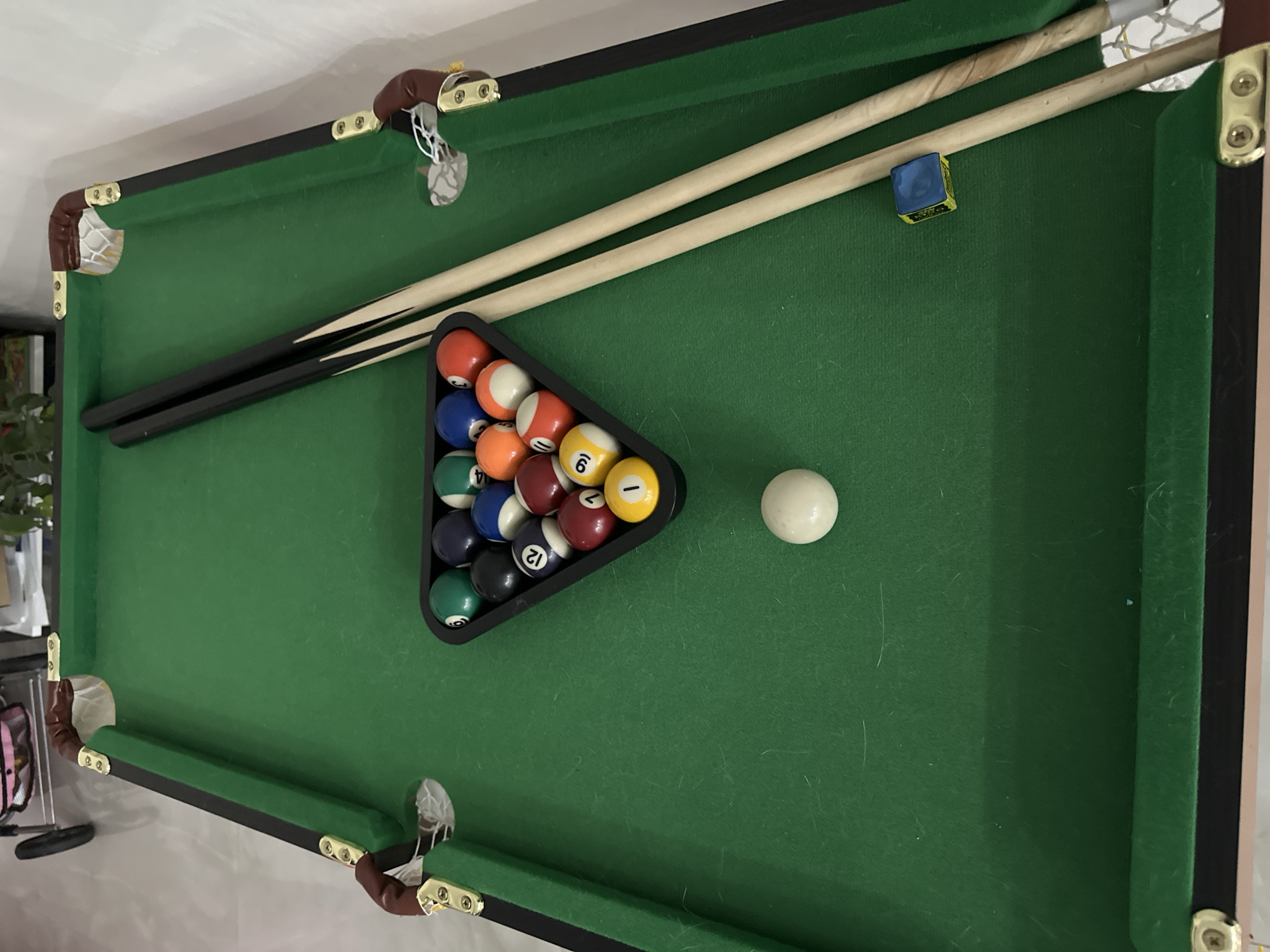 This image shows a close-up, top-down view of a green felt-covered pool table with a rack of billiard balls and two pool cues. The balls are arranged in a triangular formation, with the white cue ball positioned to the right of the rack. A blue chalk cube sits on the felt between the cues and the balls. The pool table has brown leather-like corner protectors with gold-colored fasteners. The background is blurred and appears to be a light-colored wall and possibly furniture. The lighting suggests an indoor setting, likely during the daytime, as there are no strong shadows indicating artificial light. The image is focused on the game equipment and the surface of the pool table, with no people present. The overall scene evokes a sense of leisure or preparation for a game of pool. No visible text is present on the objects in the image.