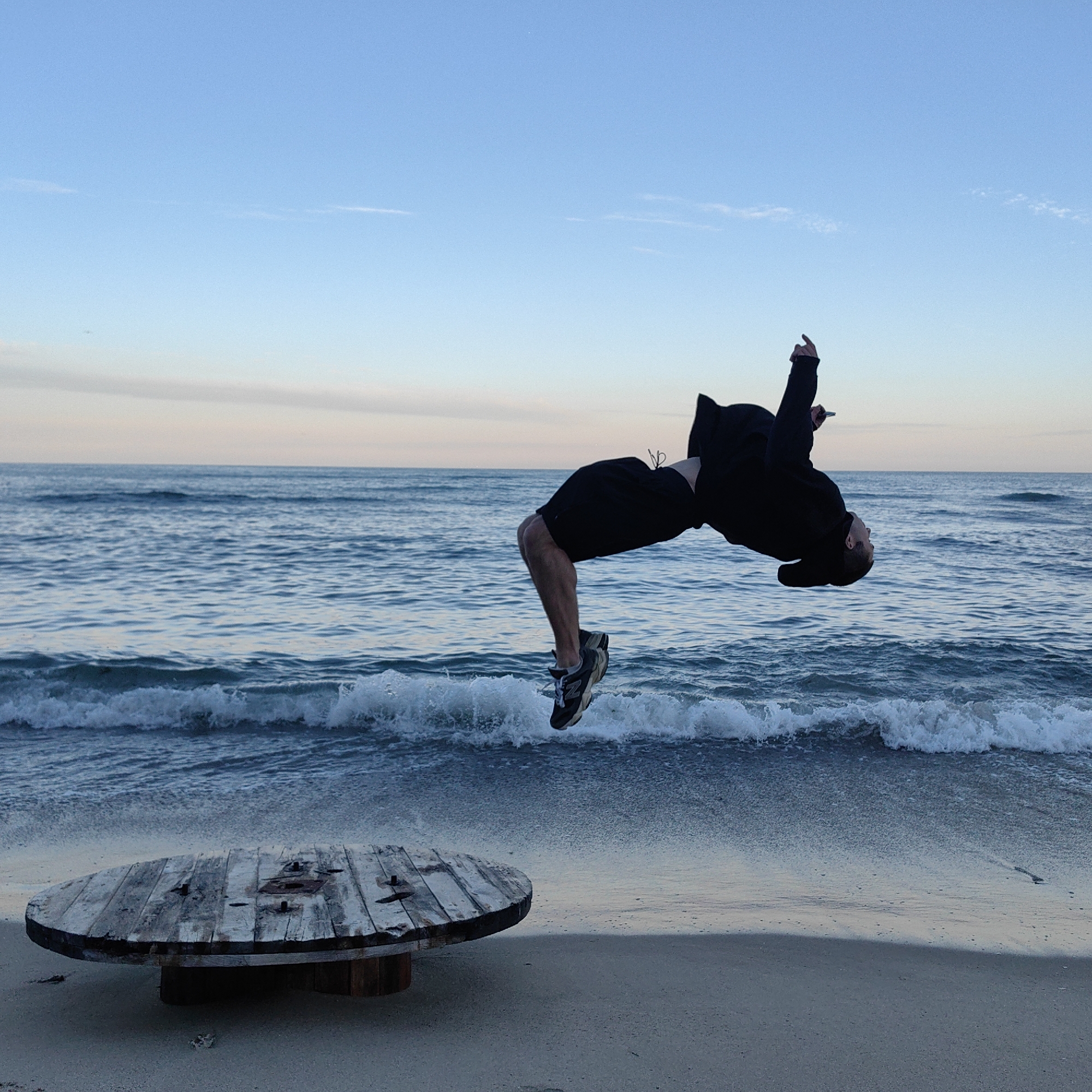 A person is performing a backflip on a beach. The person is wearing dark shorts and a dark hoodie. The background shows the ocean meeting the sky. A large wooden cable reel lies on the sand in the lower left corner. The beach is likely located on the coast of Varna, Bulgaria.