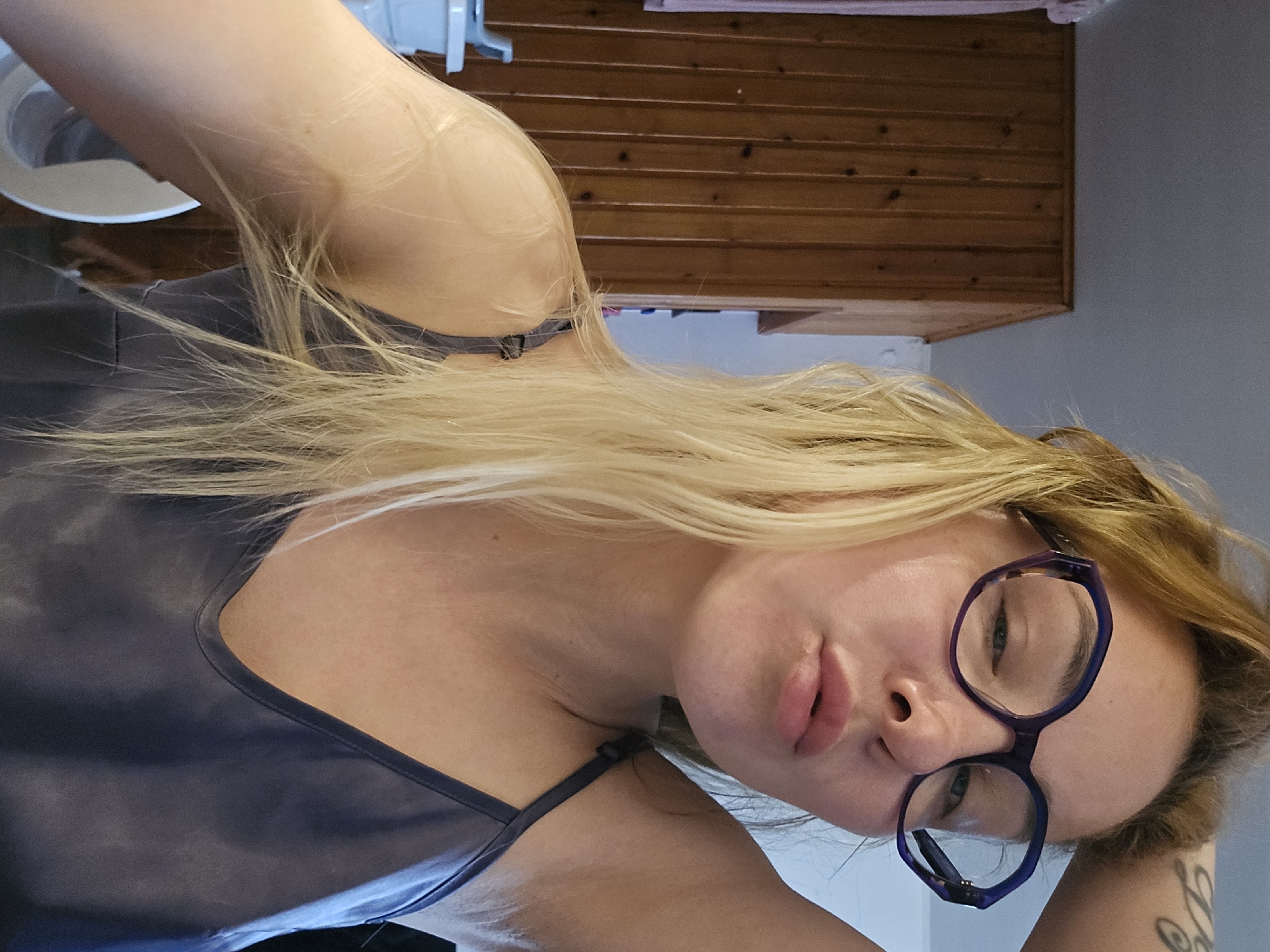A woman with blonde hair and glasses is posing for a selfie indoors. She is wearing a blue camisole. The background includes a wooden cabinet and a white sink. The presence of a sink and cabinet suggests a bathroom or kitchen setting. There is no information in the image to suggest this photo was taken in Sochi, Russia.