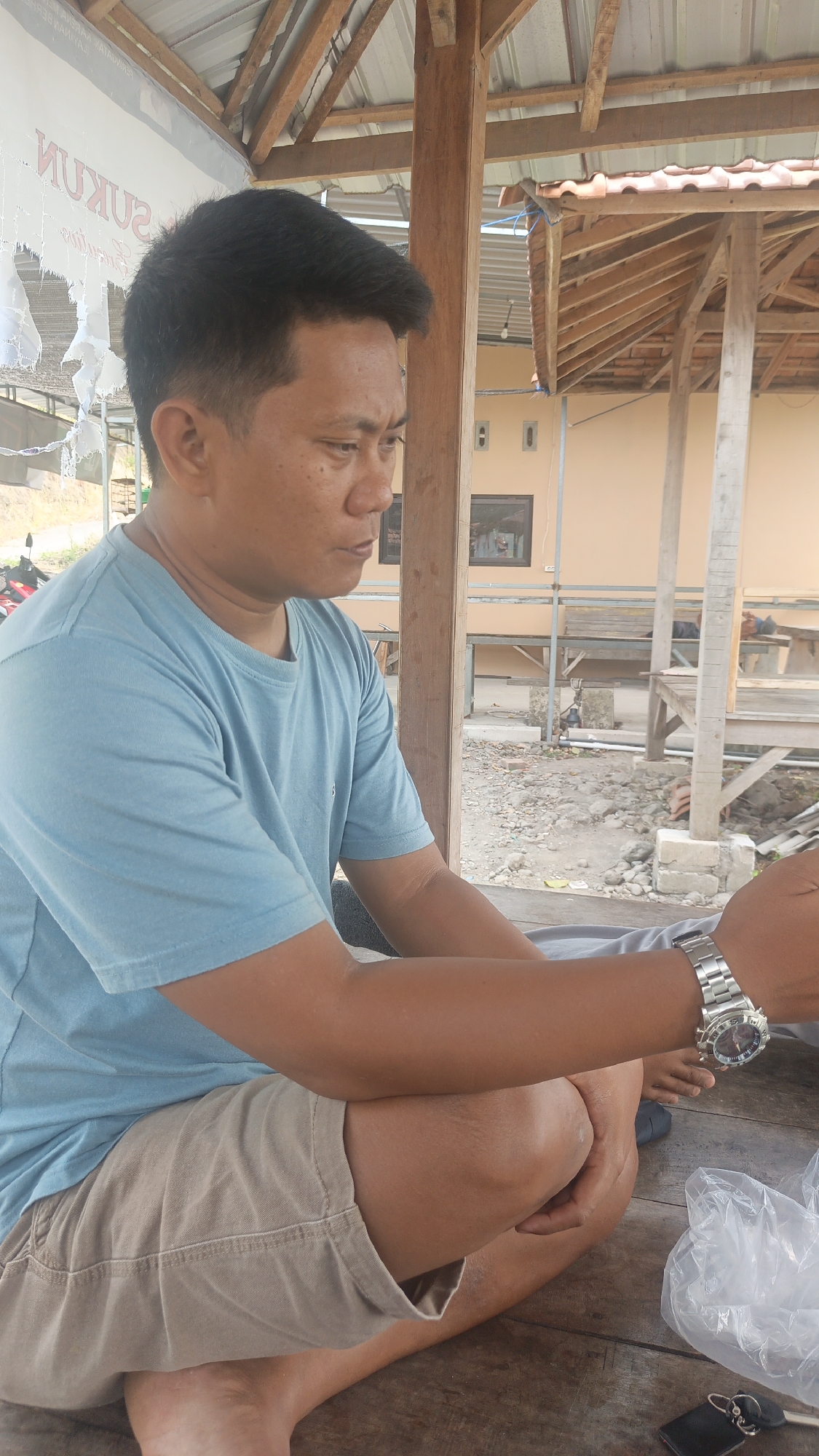 In this vertical, medium-angle shot, a man wearing a light blue t-shirt and tan shorts is seated on a wooden surface, likely a table. His body is angled to the right, with his left arm resting on his knee and his right arm extended slightly forward. He wears a silver wristwatch on his left wrist. The background depicts an outdoor setting with a partially constructed wooden structure overhead, featuring a corrugated metal roof. Behind the structure, a light-colored building with windows is visible. Scattered debris, including rocks and gravel, are present on the ground. A clear plastic bag and a set of keys are on the wooden surface in the lower right corner of the image.