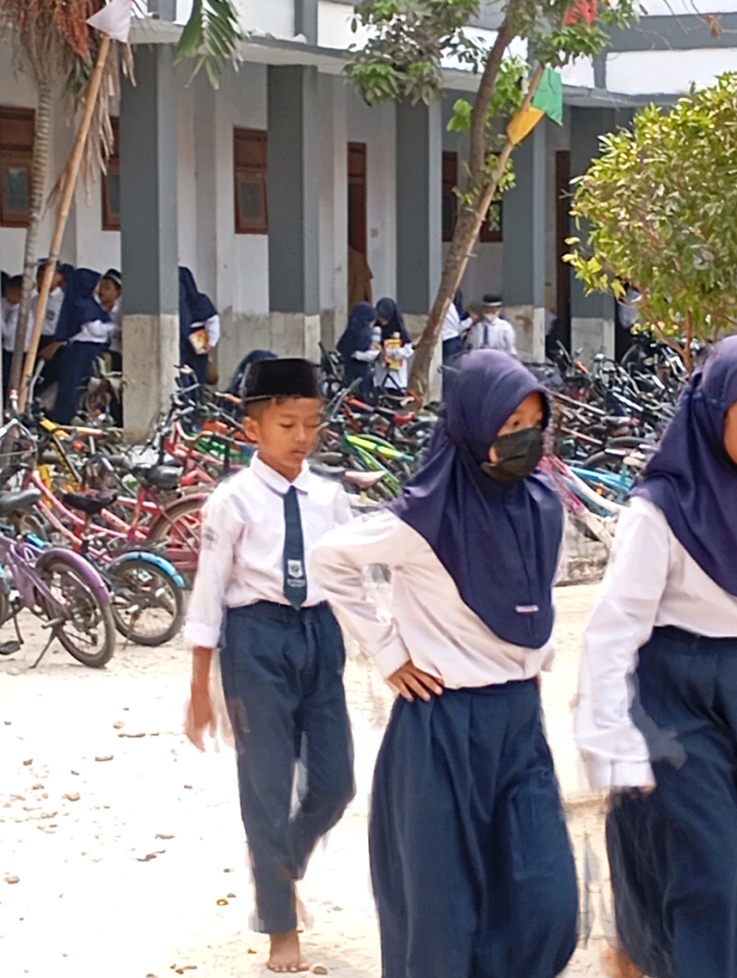 In the city of Babelan, Indonesia, a group of students in school uniforms are walking past a building with bicycles parked behind them. The students are wearing white shirts and dark-colored pants or skirts, with some wearing headscarves. One student in the foreground is a young boy wearing a black cap, white shirt, and dark pants, walking barefoot. Another student, a girl, is wearing a dark headscarf and a black face mask. The environment appears to be outdoors, possibly in a schoolyard, with a building and trees visible. The bicycles are parked in rows, suggesting a common mode of transportation for the students. The overall scene depicts a typical day at a school in Indonesia.