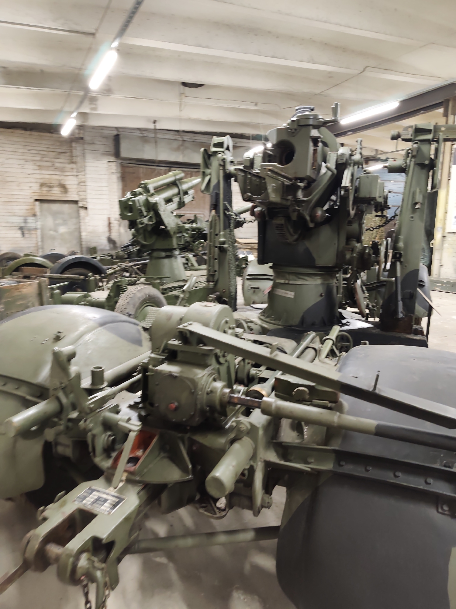 The image depicts a collection of military vehicles, specifically artillery pieces, in what appears to be a storage or workshop environment. The vehicles are primarily painted in a camouflage green color. In the foreground, a complex mechanical assembly with various gears, levers, and a tow hitch is visible. Behind this, another artillery piece with a prominent gun barrel and mounting structure is seen. Further in the background, more similar military equipment is present, some partially obscured. The environment is indoors, with fluorescent lighting illuminating the scene. The walls in the background are made of brick. The overall impression is one of military hardware being maintained or stored. The specific location in Viimsi, Estonia, is not visually evident from the image itself, but it is likely a museum or a military facility.