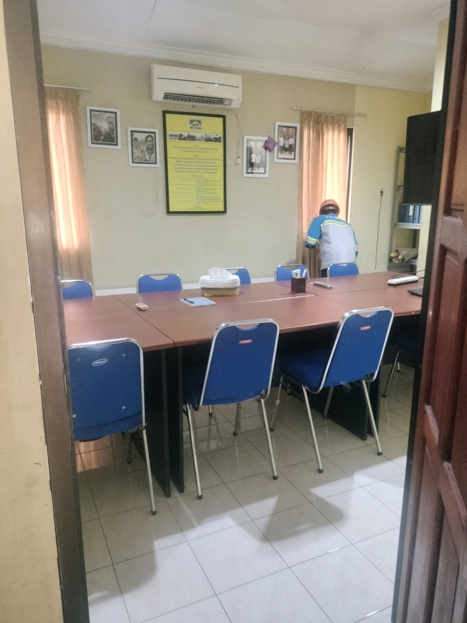 The image depicts a meeting room in Maburai, Indonesia. The room is furnished with a large, brown conference table surrounded by several blue upholstered chairs with metal frames. An air conditioning unit is mounted on the light-colored wall above the table. On the wall, there are framed pictures and a yellow poster with text. A person wearing a blue and grey work uniform and an orange hard hat is standing near the window on the right side of the room, facing away from the camera and towards the curtains. A shelf unit containing folders and other items is visible on the far right wall. The floor is tiled with a white grid pattern. The scene is viewed from the doorway of the room, with a dark brown doorframe on the right.