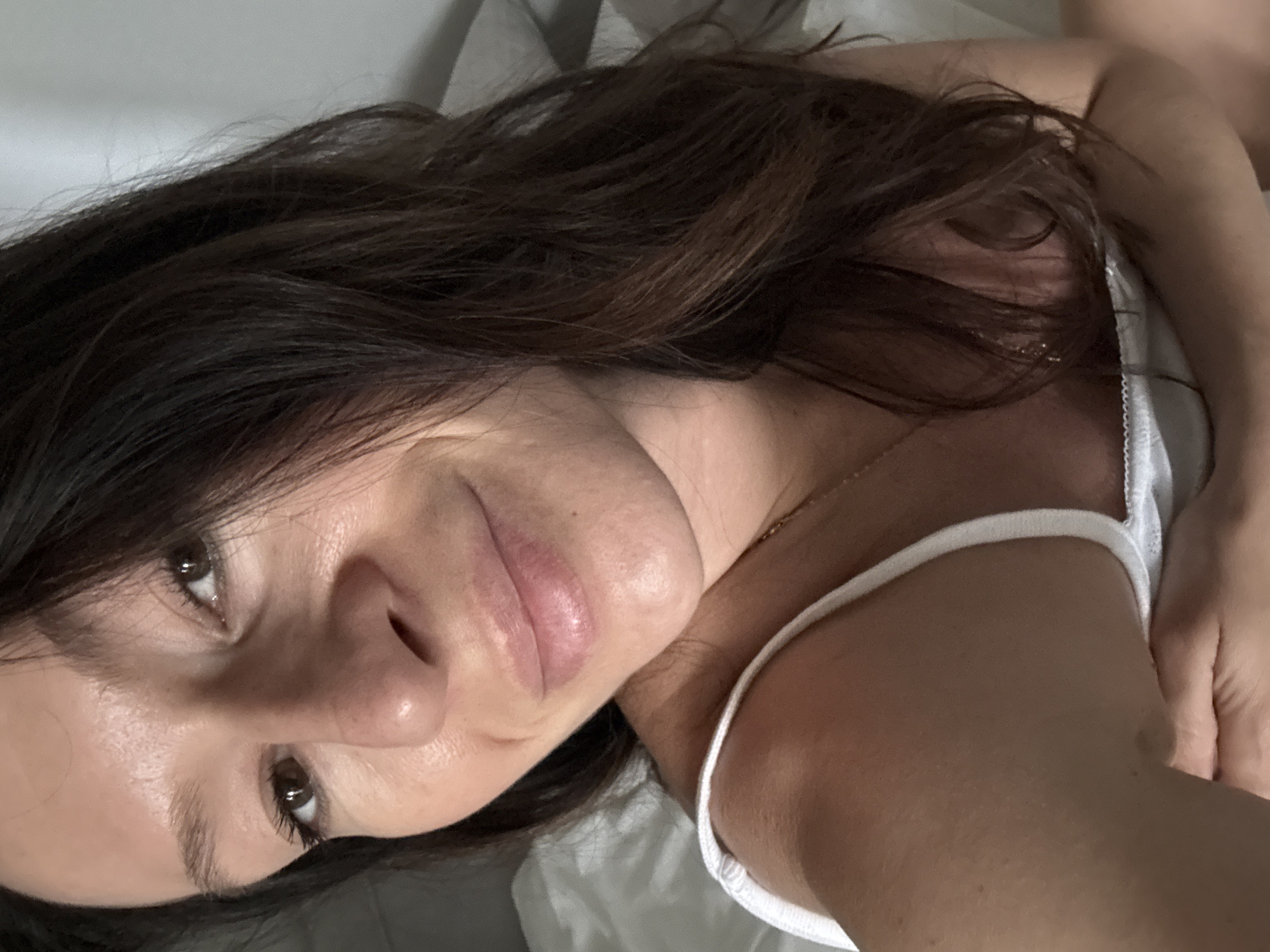 A woman with dark brown hair and brown eyes is lying on a light-colored sheet, wearing a white tank top. She is facing the camera, positioned to the left. A gold necklace is visible on her neck. No information indicates this image was taken in Chaikovsky, Russia.
