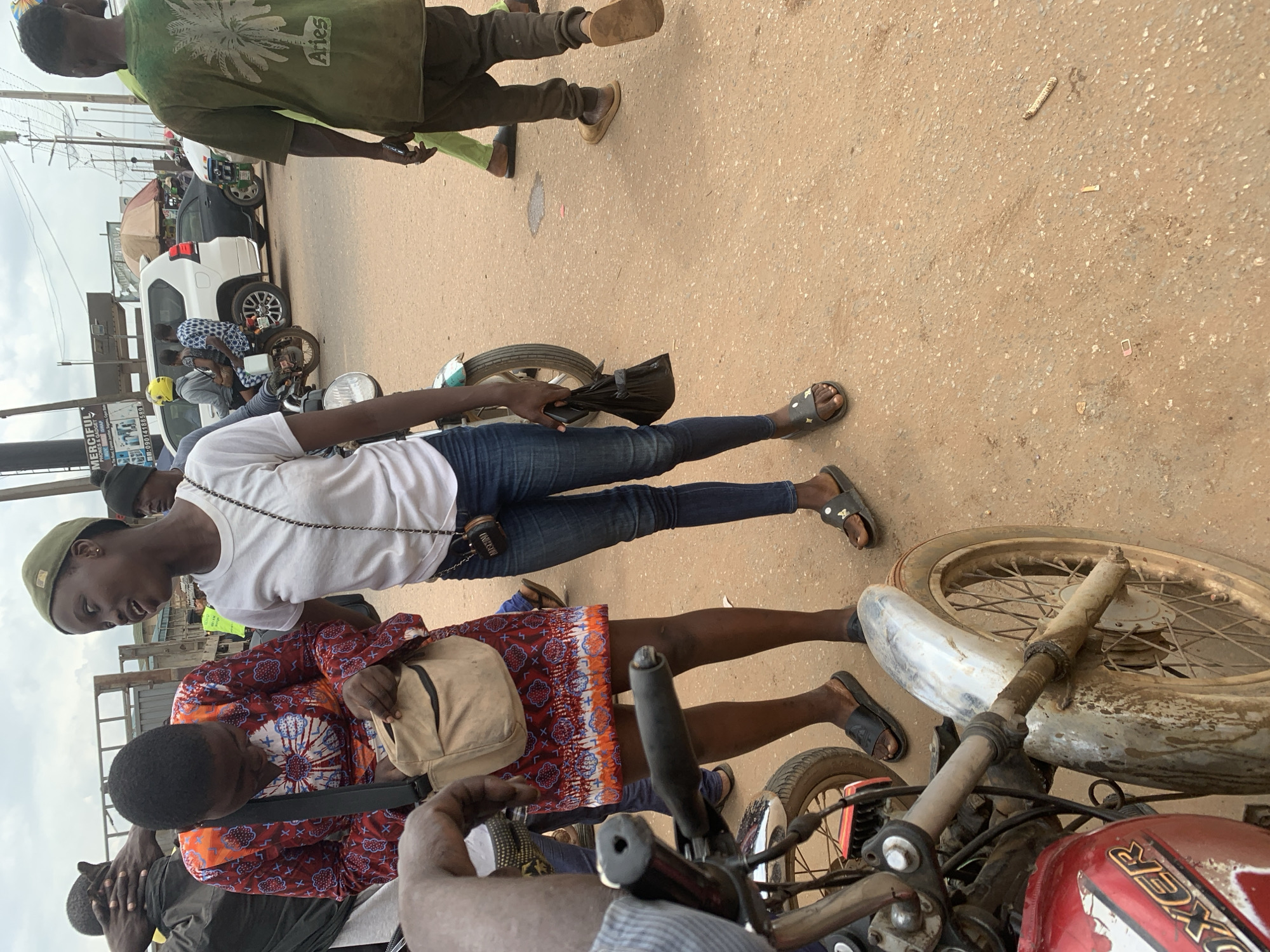 The image depicts a street scene in Shagamu, Nigeria, likely a commercial area due to the presence of various vehicles and pedestrians. The foreground is dominated by a red motorcycle, partially visible, with a muddy front fender and wheel. Two young women are the central focus. One, wearing a green cap, a white t-shirt, and blue jeans, is looking to her left and appears to be speaking. She is holding a small black bag in her right hand. The other woman, in a red and white patterned dress and a beige cross-body bag, is looking down and appears to be rummaging in her bag. Both women are wearing dark colored sandals.
In the background, other individuals are visible, including a man in a green t-shirt with a palm tree graphic and the word "Aries" on the back, walking away from the camera. Further back, traffic is present, including a white SUV and several motorcycles. A sign with text partially obscured by a tree can be seen, suggesting a commercial establishment. The ground is paved with a sandy, reddish-brown material, typical of many Nigerian roads. The sky is overcast. The overall atmosphere suggests a busy, everyday street environment.
