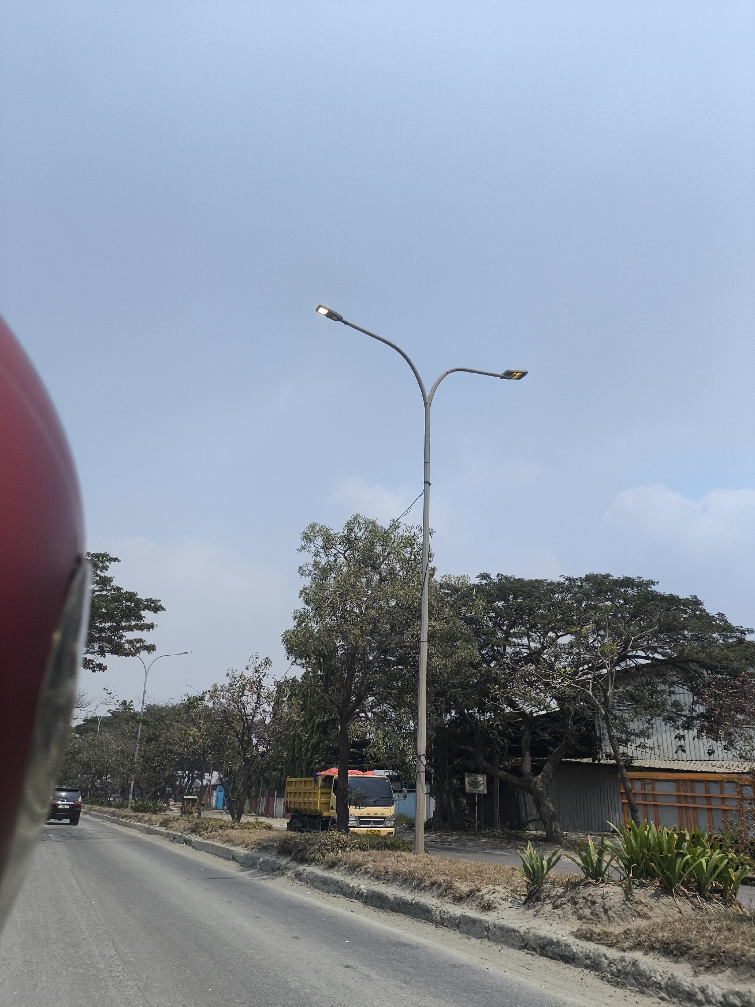 The image depicts a street scene in Cilegon, Indonesia, under a pale blue sky with scattered clouds. A prominent double-headed street lamp stands on the right side of the road, with one light illuminated. The road itself is paved, with a sandy verge and curb to the right.
On the roadside, several trees of varying sizes are visible, some with dense foliage and others with bare branches. A yellow truck with a dumper bed is parked on the verge, partially obscured by trees. Behind the truck, a building with a metal facade and wooden accents can be seen. In the distance, more trees and structures line the street.
A dark-colored vehicle is driving away from the camera on the left side of the road. The foreground is dominated by the out-of-focus curve of a red object, likely a helmet, suggesting the photo was taken from a moving vehicle. There are no visible people in the scene. The overall impression is of an industrial or commercial area on a sunny day.