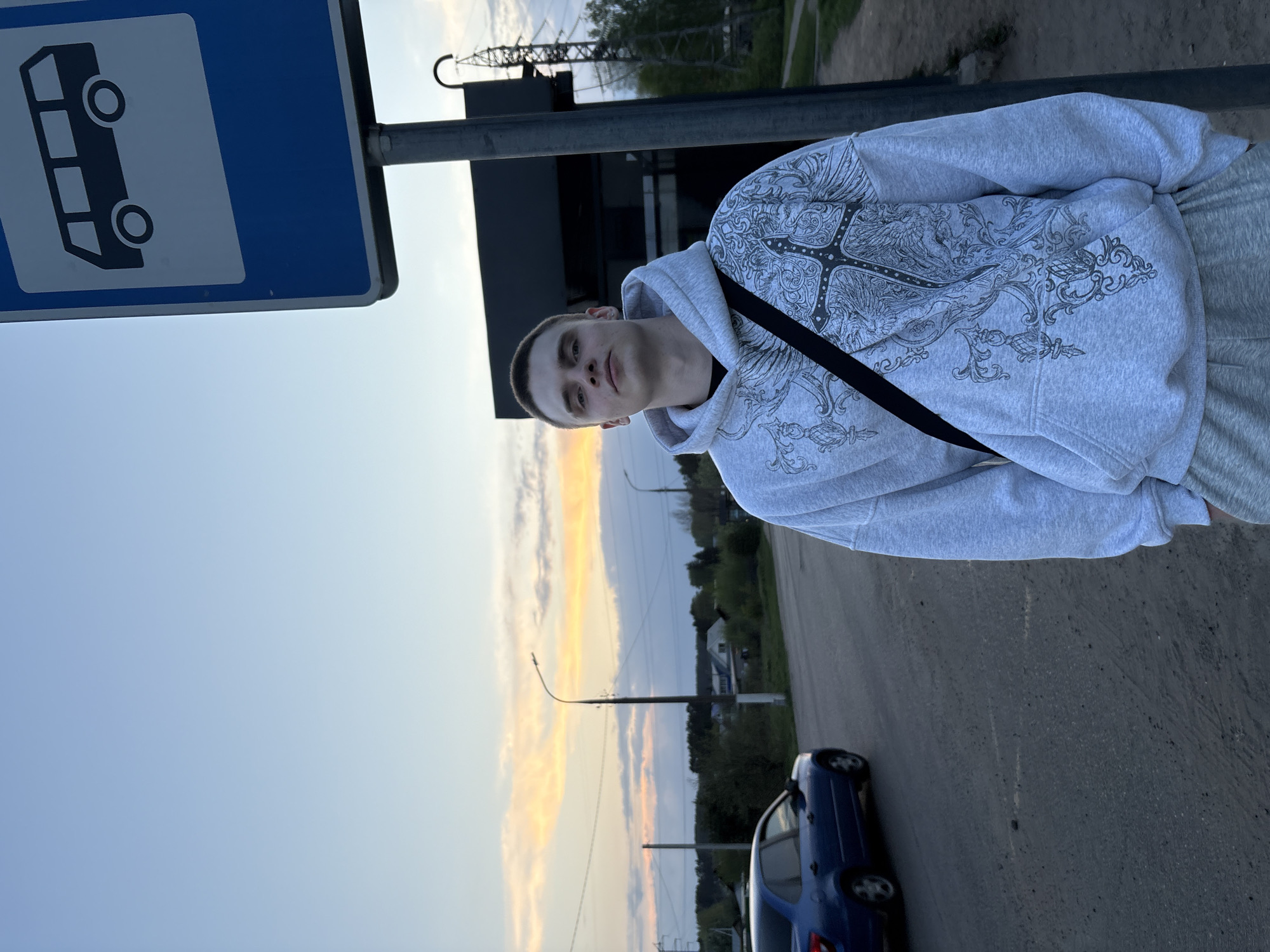 The image shows a young man in a gray hoodie standing near a bus stop sign. He has short hair and a black strap across his chest. Behind him is a road with a blue car parked on the side. In the distance, there are trees, power lines, and a partially visible building. The sky is a mix of blue and orange, suggesting it's either sunrise or sunset. This scene is in Chaikovsky, Russia, as indicated by the bus stop sign design.